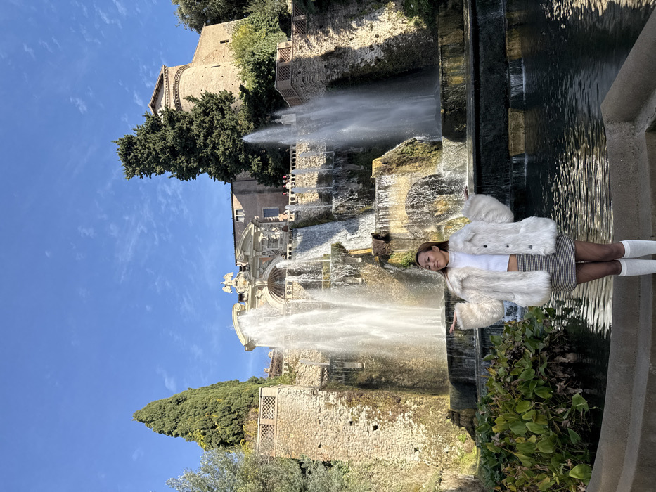 An outdoor, brightly lit scene at the Villa d'Este in Tivoli, Italy, features a multi-tiered fountain complex. A single East Asian female subject, estimated to be in her 20s or 30s, stands in the foreground, positioned slightly to the right of the central axis. She has long, straight brown hair and is wearing a white faux fur jacket over a white cropped long-sleeved top, a black and white plaid mini-skirt, dark sheer tights, and white knee-high socks. Her arms are outstretched to the sides with palms facing upwards, and her head is tilted slightly right with a faint smile, looking towards the upper right.
Behind the subject, the expansive Fountain of Neptune cascades water over multiple stone tiers and pools. The central upper portion of the fountain exhibits ornate Baroque architecture with decorative elements, including a prominent crest. Tall, dark green cypress trees flank the fountain structure, one on the far left and another to the right. A group of indistinct people are visible on a stone walkway behind the fountain, near the center-right.
In the background, above the fountain, the light-brown stone facade of the Palazzo d'Este is visible, including a distinct rounded tower on the far right. The sky is clear and bright blue with minimal scattered white clouds. The scene is illuminated by natural sunlight, casting shadows primarily towards the right side of the structures.