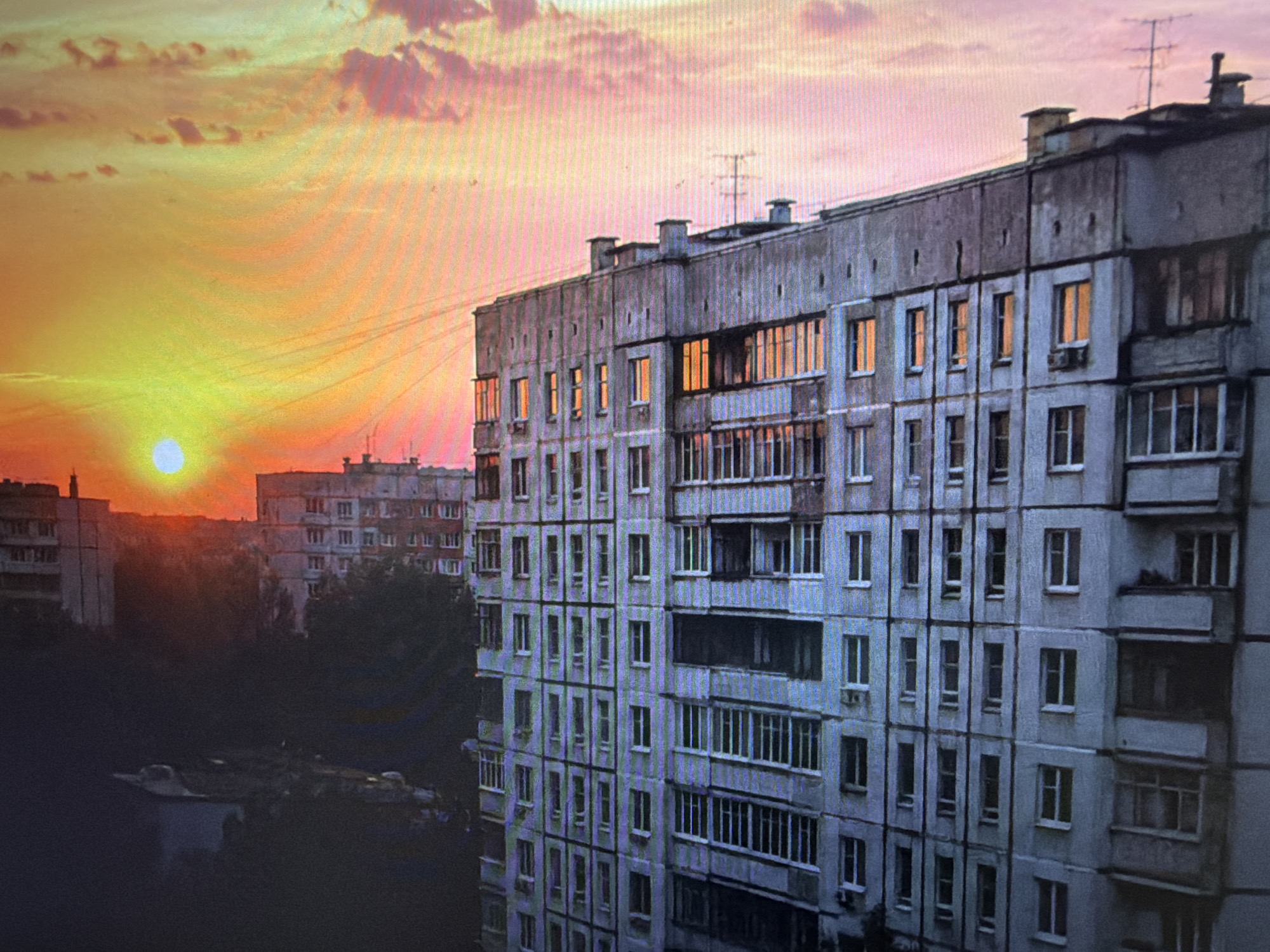 The image depicts a residential apartment building in an urban setting at sunset. The sky is painted with warm hues of orange and pink, with the sun visible as a bright disc on the left side. The building, a typical Soviet-era concrete structure, is grey and shows signs of wear. It is a multi-story building with numerous windows, many of which are illuminated from within, suggesting occupancy. Balconies with railings are visible on some floors. In the foreground, dark silhouettes of trees and other structures are present, partially obscuring the lower parts of the building. The overall atmosphere is one of urban life at the end of the day, with the natural beauty of the sunset contrasting with the utilitarian architecture of the city.