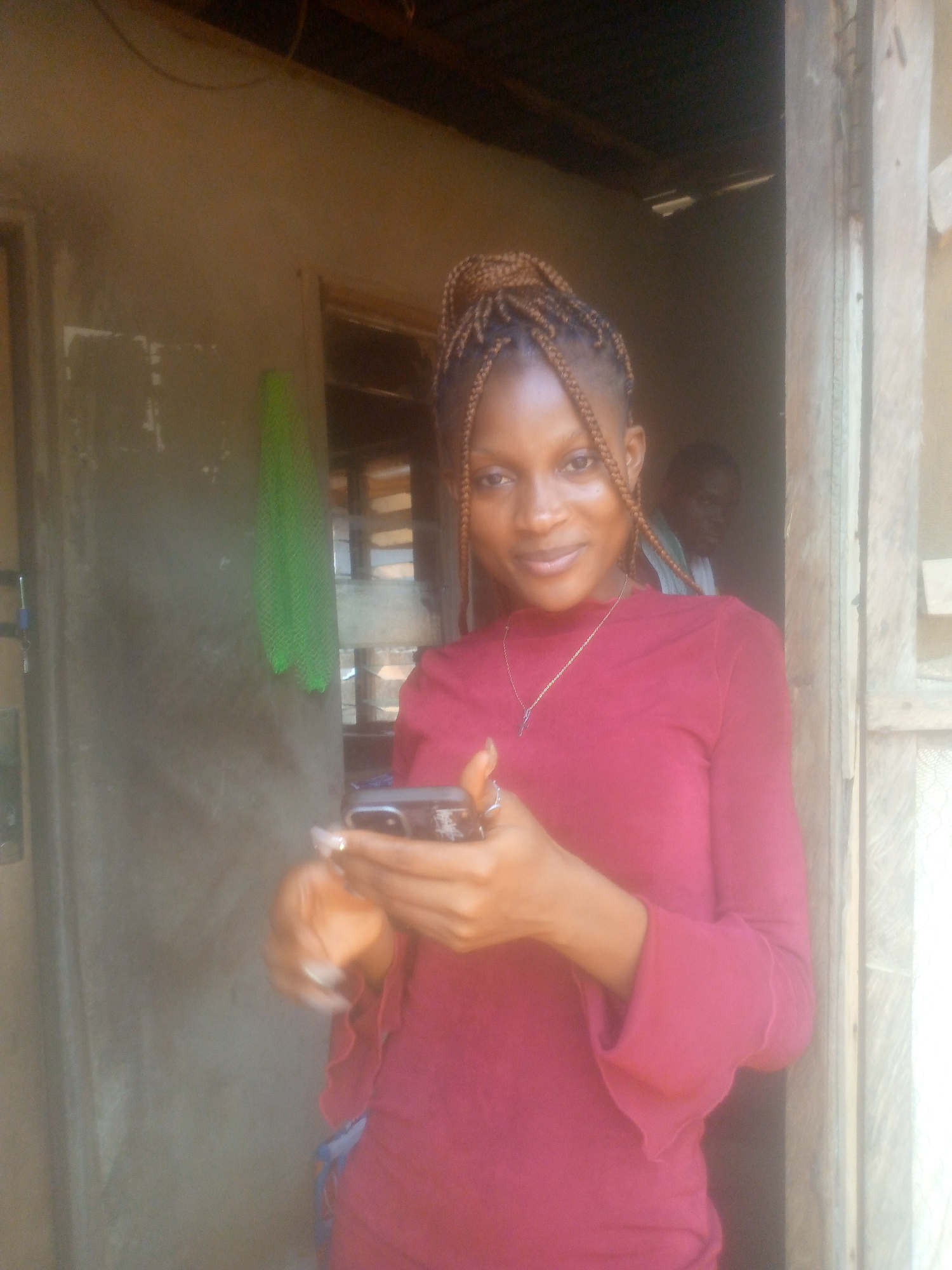 A young woman with braided hair stands in a doorway, looking at the camera and holding a smartphone. She is wearing a long-sleeved, red-purple top and a thin necklace. Behind her and partially visible through the doorway is a male figure wearing a light-colored top. The environment appears to be a simple, possibly rural, building with a green towel hanging on the wall to the left. The location is given as Unknown, Unknown.