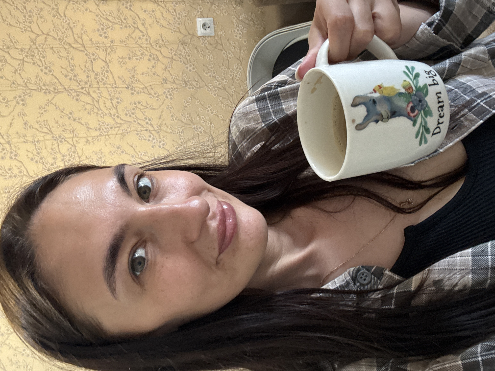A woman with long, dark hair and fair skin is holding a mug with the words "Dream big" printed on it. The mug also features a graphic of a gray dog. She is wearing a black shirt and a gray plaid jacket. The background shows a yellow wallpaper with a floral design and an electrical outlet. The location is not identified in the image. It is not possible to determine if the image was taken in the city of Sochi, Russia.