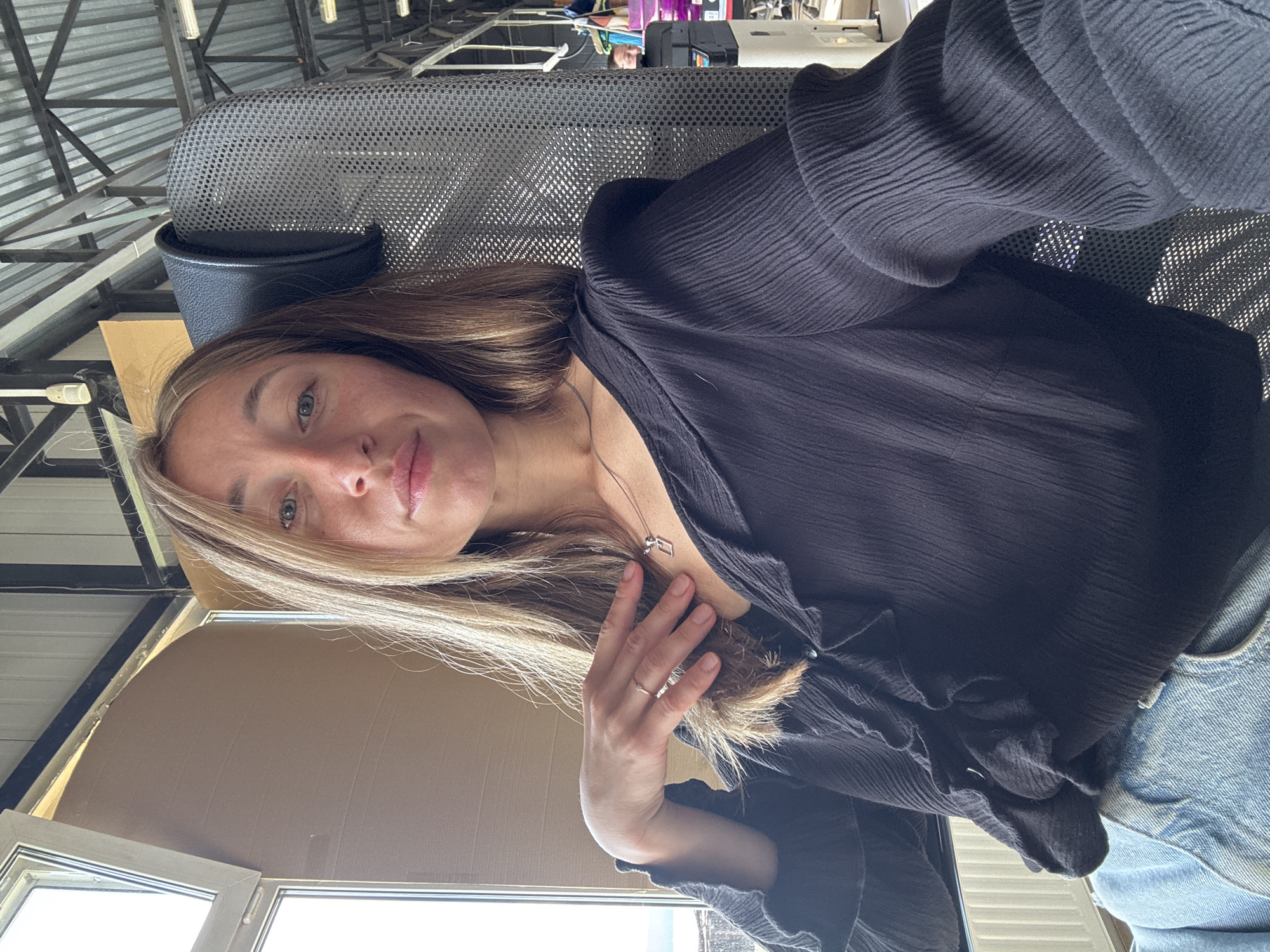 A woman with long, light brown hair is seated in a chair, taking a selfie. She is wearing a black blouse with ruffles and a silver necklace with a square pendant. Her left hand is resting on her chest, and she is wearing a ring on her ring finger. She is looking at the camera with a neutral expression. The background appears to be an indoor space with a corrugated metal ceiling and large windows. The chair she is sitting in has a mesh back and headrest. Based on the architecture visible, this could be a building associated with industrial or public transportation infrastructure, such as a bus or train station. Without more specific visual cues, pinpointing the exact location in Chaikovsky, Russia, is not possible. However, the overall impression is of a functional, perhaps utilitarian, indoor environment.