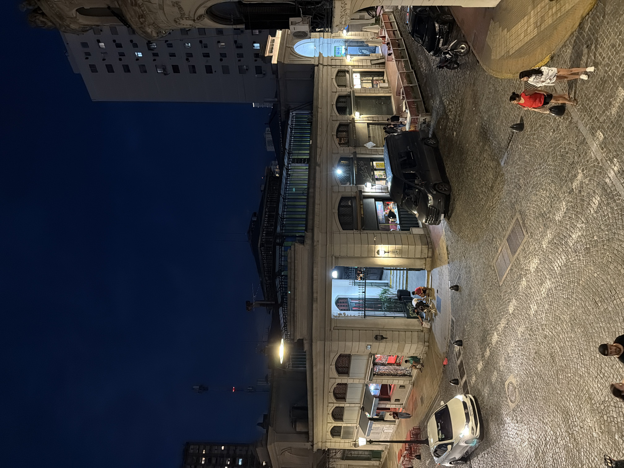 This is an outdoor scene taken at night in what appears to be a European-style street in Buenos Aires, Argentina. The street is paved with cobblestones. Buildings line the left side of the street, illuminated by street lights and lights from within the structures. The building facades are ornate, and some have awnings over what appear to be storefronts. People are walking on the right side of the street, and cars are parked along the building side. There's a white car in the foreground. Above the buildings, the night sky is a deep blue. The angle of the picture is somewhat tilted.