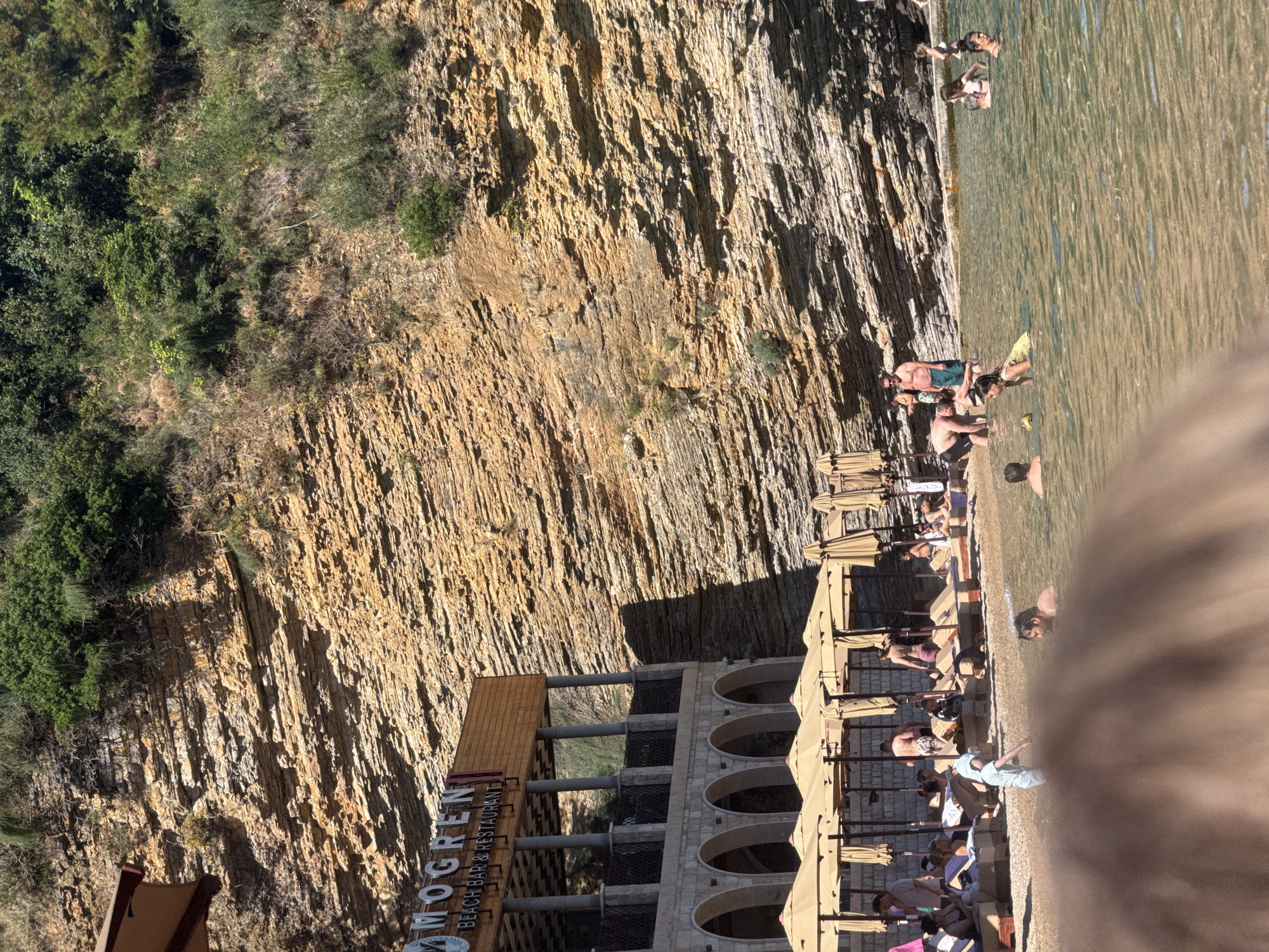 This image shows a beach scene with people enjoying the water and sun. The beach is situated at the base of a steep, rocky cliff face covered in sparse vegetation. The water is clear and calm, with ripples indicating shallow depth. Several umbrellas are set up on the beach, providing shade for beachgoers who are relaxing on lounge chairs or in the sand.
People are engaged in various activities, including swimming, wading, and sunbathing. Some are standing in the water, while others are seated or lying down on the shore. The overall atmosphere appears relaxed and leisurely.
A building with arches is visible on the right side of the image, with text above it that reads "MOGREN BEACH BAR & RESTAURANT". This indicates the location as Mogren Beach in Budva, Montenegro. The time of day appears to be sunny and warm, likely midday or afternoon, given the bright sunlight casting shadows. The clear sky and calm water suggest pleasant weather. The scene conveys a sense of vacation and enjoyment.