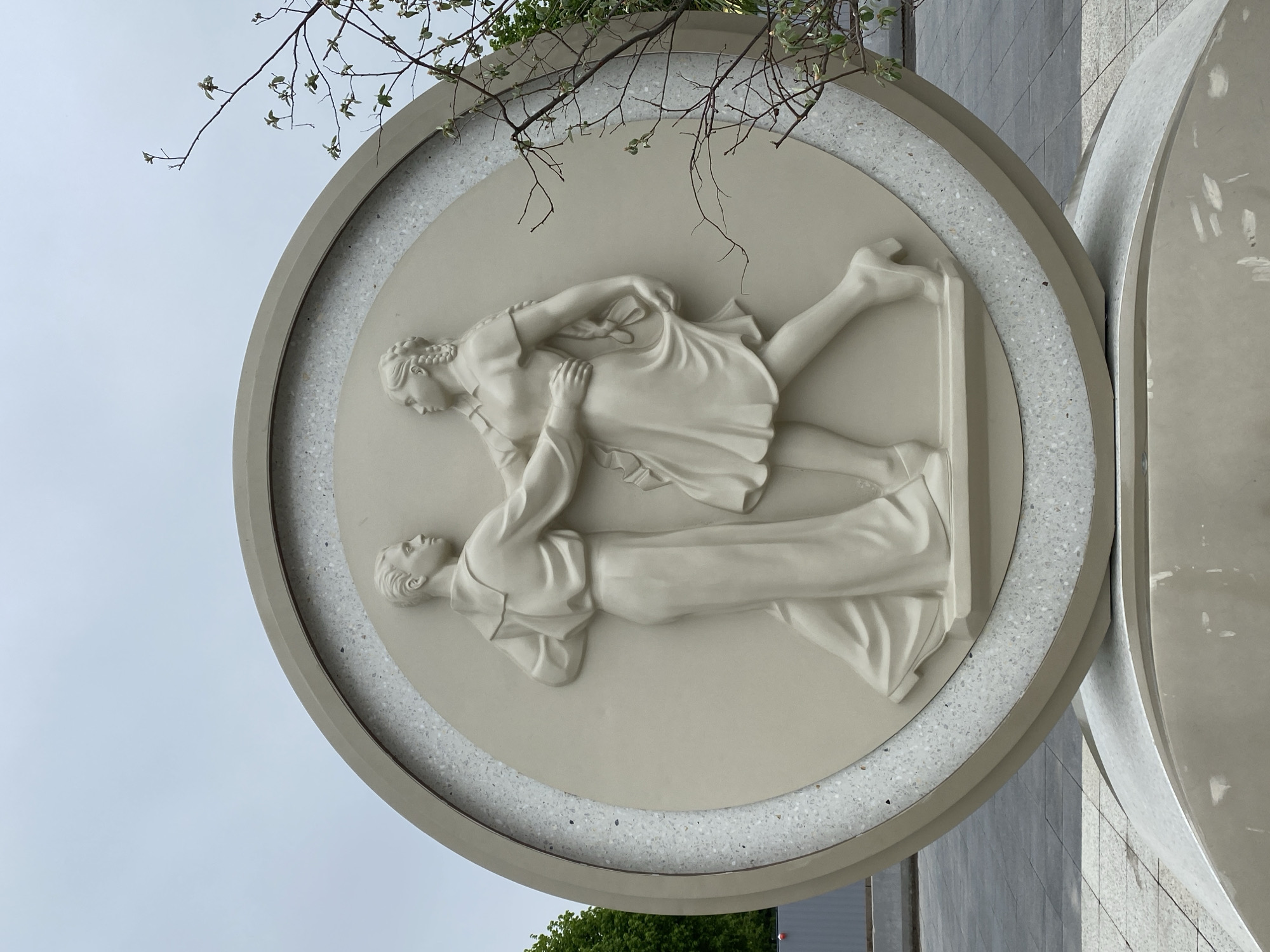 A stone bas-relief sculpture is depicted in a close-up shot. The sculpture, contained within a circular frame with a speckled stone border, portrays two figures in classical attire in a dynamic, intertwined pose. One figure appears to be supporting or catching the other, who is positioned higher. The scene is partially obscured by tree branches with sparse foliage. The sculpture is part of a larger structure, as evidenced by a portion of another circular element visible on the right. The background suggests an urban setting with architectural elements and foliage. The location is Unknown, Unknown.