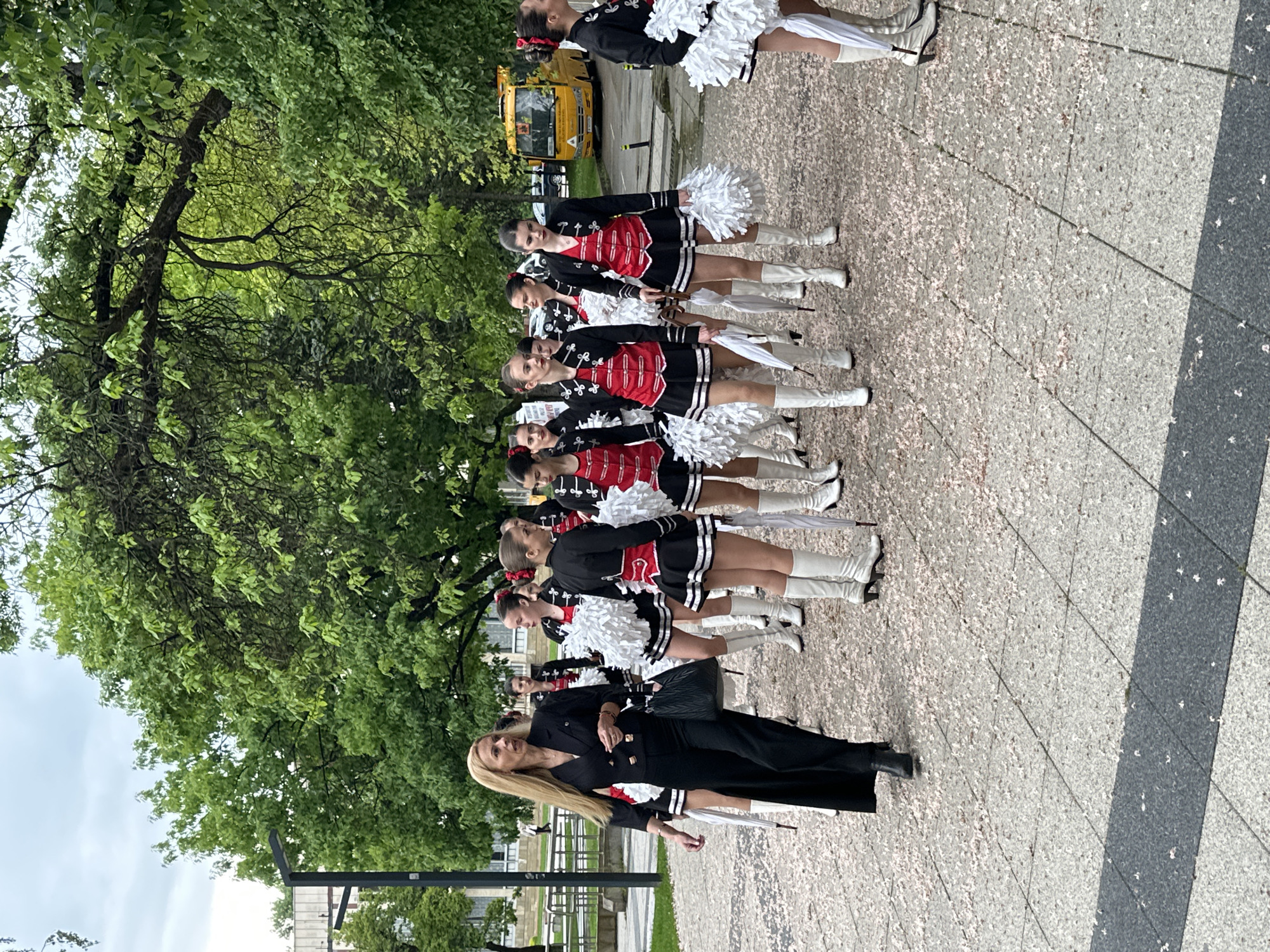 A line of women in marching band uniforms stand on a patterned stone surface near trees in Montana, Bulgaria. The women are wearing black and red jackets, white boots, and some are holding white pom-poms. A woman in a black suit stands at the front of the line. A yellow truck is visible in the background. The sky is overcast.