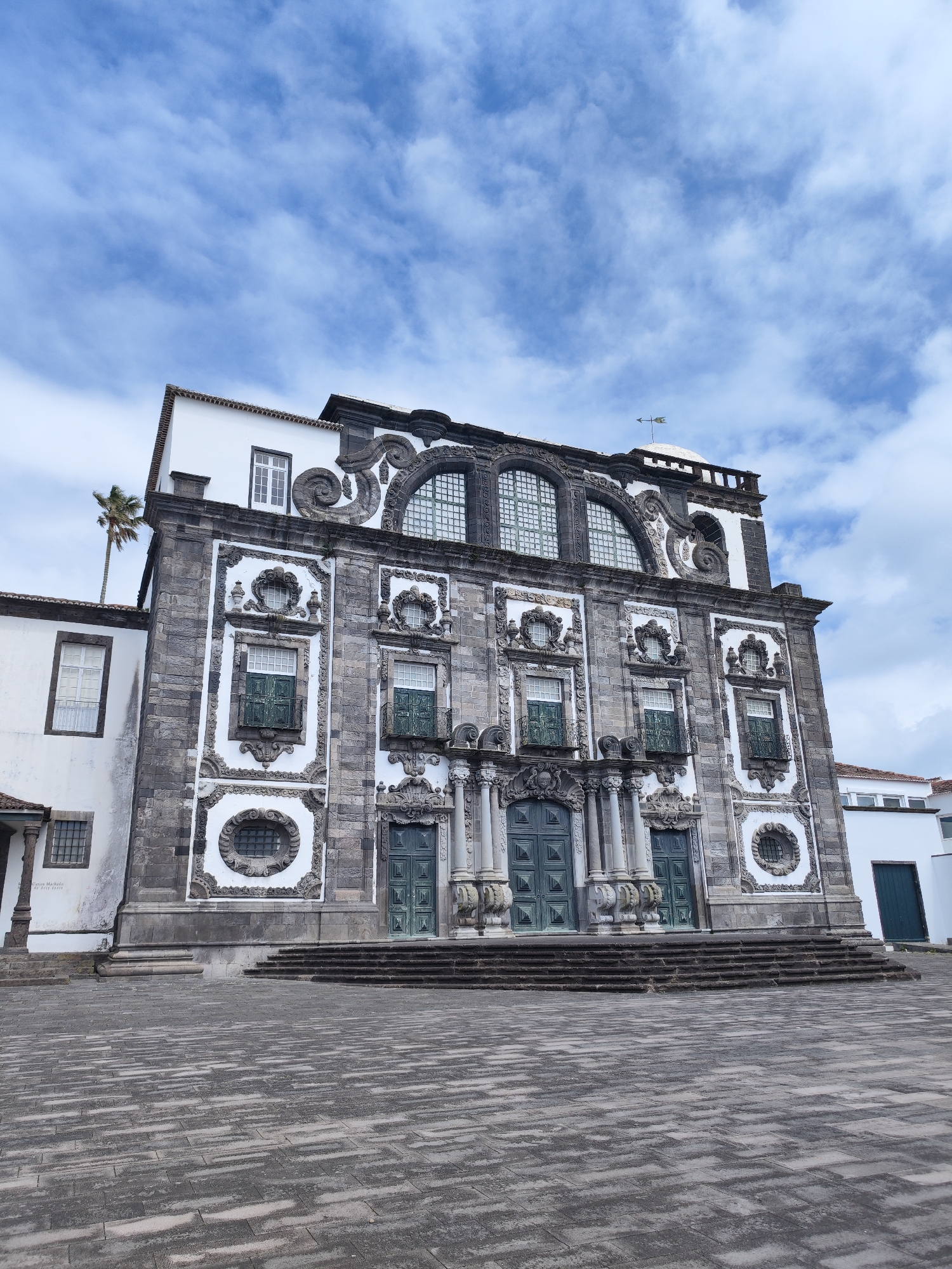 The image displays the grand and ornate facade of a historic building, likely a church, under a bright, partly cloudy blue sky. The structure features a distinctive Baroque or Mannerist architectural style, with a base of dark grey stone blocks contrasted by white plastered sections richly adorned with decorative carvings, scrolls, and sculptural elements.
At the ground level, three prominent, dark green double doors serve as the main entrances, each flanked by decorative columns and elaborate stone pediments. These doors are accessed by a wide series of shallow, dark grey stone steps. Above the entrances, the mid-section of the building showcases multiple rectangular windows with intricate iron railings, set within highly embellished surrounds. The upper story is characterized by large, multi-paned arched windows, framed by extensive stonework and large decorative scrolls, leading up to a detailed cornice and a flat roofline. A small dome topped with a weathervane is visible on the right side of the roof.
To the left of the main facade, a simpler white building adjoins the structure, with a tall palm tree visible peaking out from behind it. The foreground consists of an expansive public square, paved with an irregular pattern of light and dark grey stone blocks, showing signs of wear and staining. No people or activities are visible, contributing to a serene and still atmosphere.
The lighting indicates it is daytime, likely late morning or early afternoon. On the white wall of the adjacent building to the left, below a window, faint black text is discernible, reading "MARIANO MACHADO" and "R. AGOSTINHO DE ORNELAS". The architectural style, the palm tree, and the general appearance are consistent with the context of Ponta Delgada, Portugal.