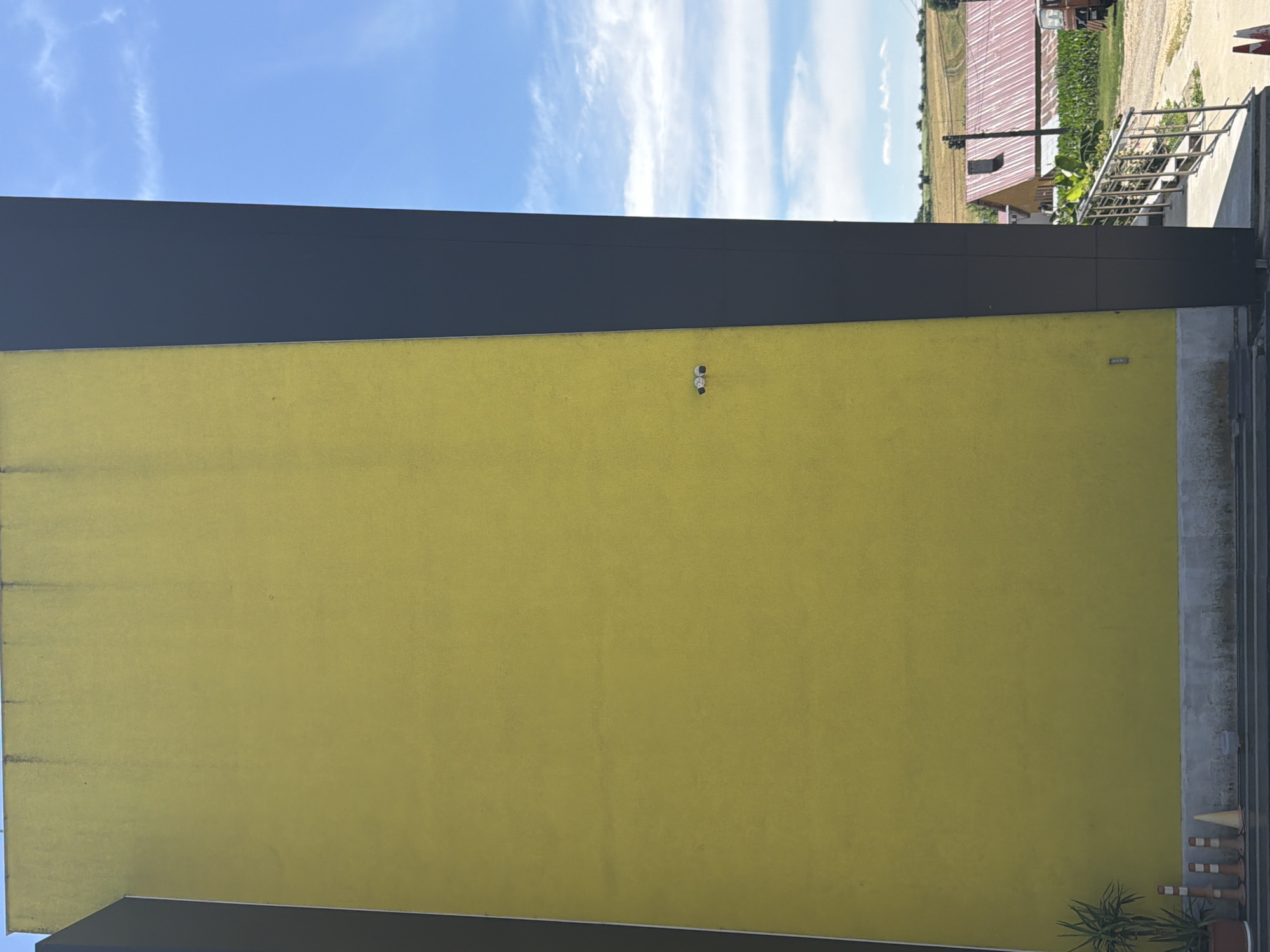 This is a vertical, medium-shot, eye-level photo of a bright yellow stucco wall with a dark gray overhang at the top. The wall occupies most of the frame, with the sky and a rural landscape visible above the overhang. The sky is a vibrant blue with scattered white clouds, suggesting a clear, sunny day. The time of day appears to be daytime, likely late morning or early afternoon, given the brightness of the light.
On the yellow wall, two small, white security cameras are mounted. The overall scene is one of an outdoor setting, possibly a building with an adjacent outdoor area. The texture of the stucco is visible, showing slight imperfections and some darker streaks or smudges, particularly along the left edge and top.
In the background, beyond the overhang, a portion of a rural landscape is visible. This includes a large building with a red-ribbed metal roof, possibly a barn or agricultural structure, and fields of crops. A truck or vehicle is partially visible near the red-roofed building. A metal railing or fence is also present in the background, leading towards the building. The location is identified as Dolno Ablanovo, Bulgaria. There is no text visible in the image. The overall impression is a mundane, daytime scene of an exterior wall and its surroundings.