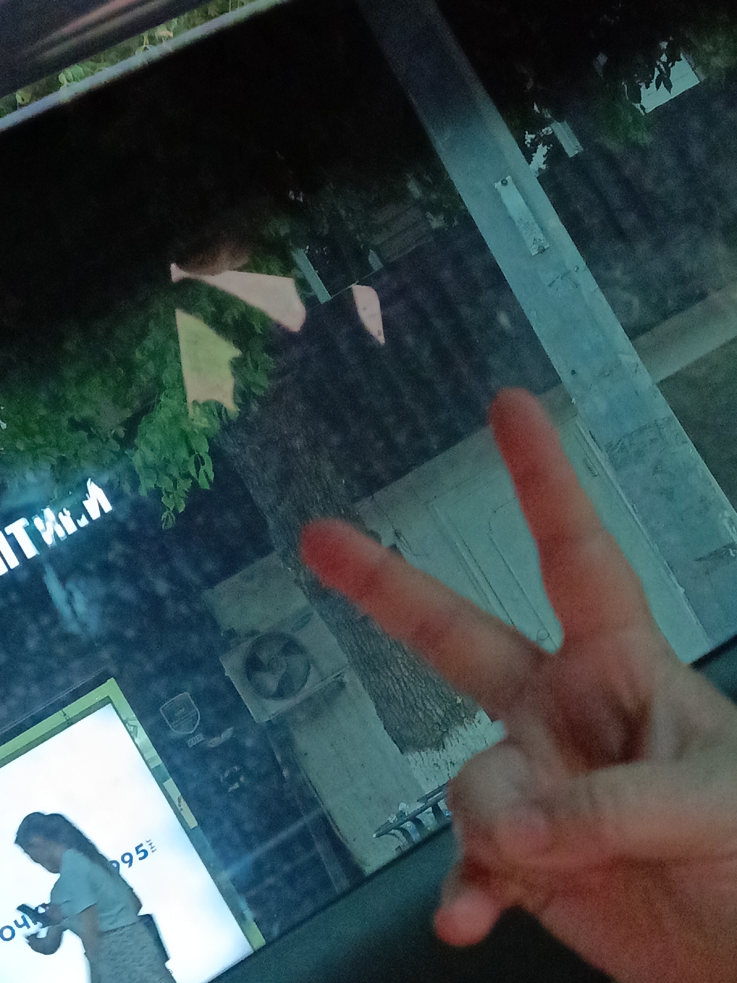 The image, captured on a vertical orientation, depicts a nighttime scene, likely in Shymkent, Kazakhstan, judging by the visible signage. In the foreground, a hand is held up, forming a peace sign with its index and middle fingers extended. The hand is illuminated by an orangish-red light source, giving it a warm hue.
In the background, a neon sign with white lettering that appears to read "ITVM.И" glows against the darkness, suggesting a commercial establishment. To the left of this sign, a large, illuminated display showcases a silhouette of a woman holding a phone, with partial text visible, including "095". This suggests an advertisement for a telecommunications service or a product related to mobile technology.
The environment is reflected in what appears to be a window, possibly from a vehicle, as indicated by the framing and the overall perspective. This reflection reveals a dimly lit street scene with trees and a building facade. An outdoor air conditioning unit is visible on the wall of the building. The lighting is predominantly from artificial sources, contributing to the nocturnal atmosphere. The overall impression is that of an urban setting at night, viewed through a reflective surface.