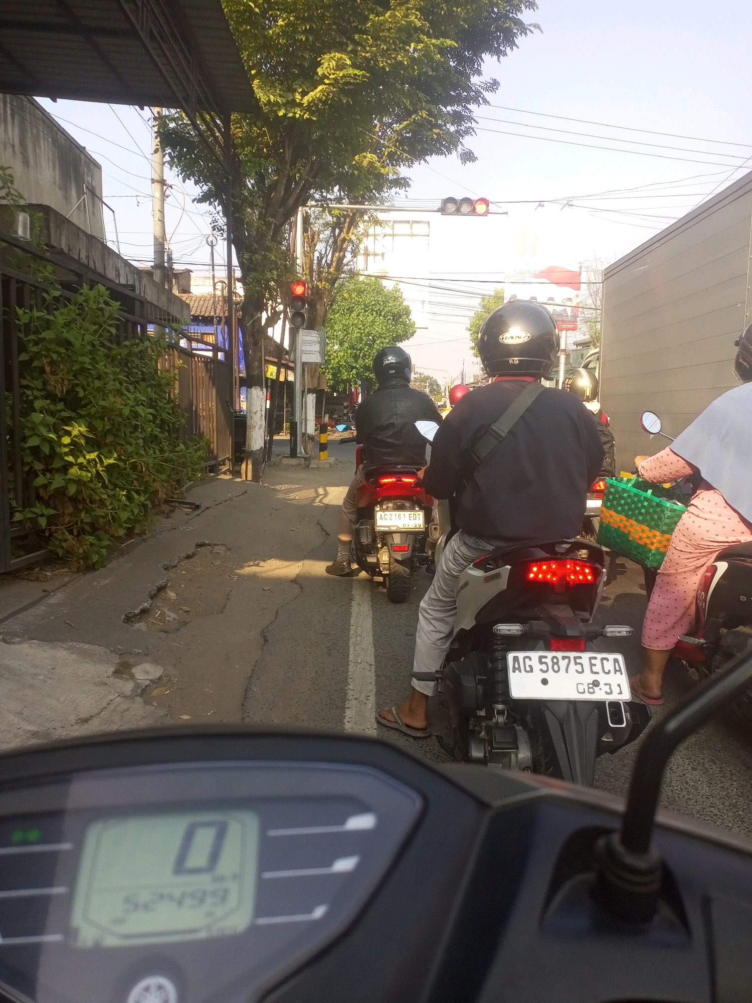 The image shows a traffic scene with multiple motorbikes in a city identified as Gurah, Indonesia. The perspective is from the point of view of a rider looking forward, with their motorbike's instrument panel visible in the foreground. Several motorbikes are stopped in traffic, indicated by their brake lights. Two riders are clearly visible, one in the immediate foreground and another slightly ahead. The rider in the foreground is wearing a black helmet and a dark long-sleeved shirt with grey pants and flip-flops. The rider ahead is wearing a black helmet and a black leather jacket. Another rider, a woman, is on a motorbike to the right, wearing a pink patterned outfit and carrying a green and yellow woven bag. The environment includes a paved road with some cracks and potholes, a sidewalk with lush green foliage, and buildings on the left side. Traffic lights are visible in the distance, with the red light illuminated. The sky is clear and blue. The time of day appears to be late afternoon or early evening, given the warm lighting. The license plate of the motorbike in the foreground reads "AG 5875 ECA".