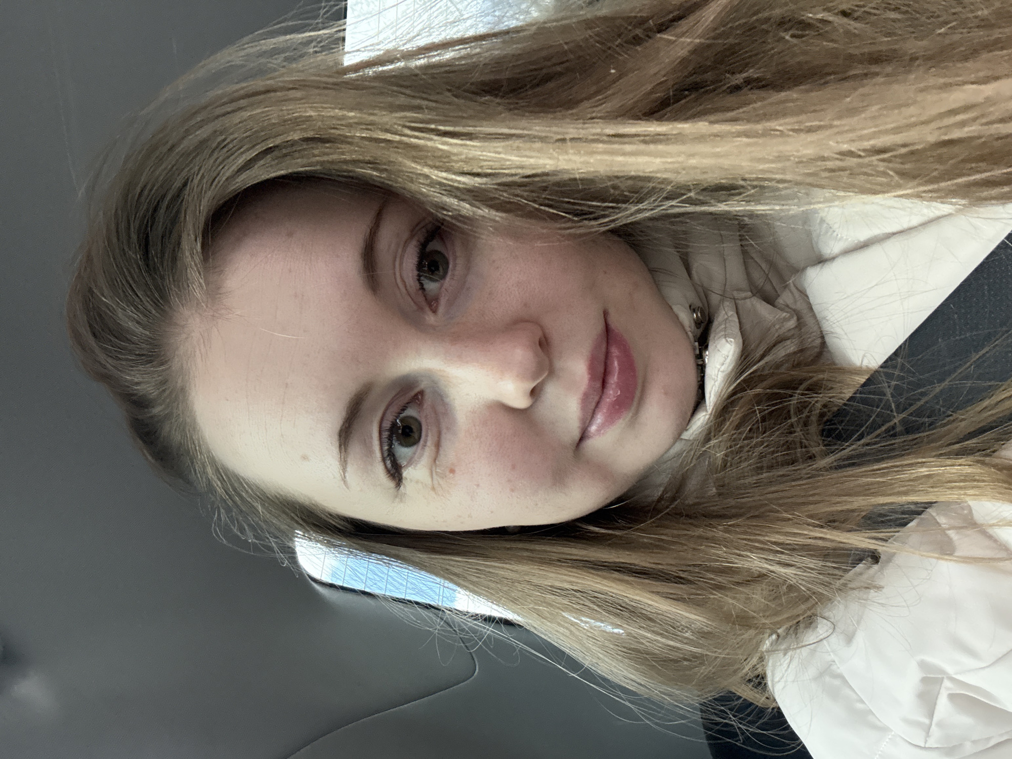 A young woman is captured in a close-up selfie, taken from a slightly upward angle, with her head tilted, resulting in a rotated composition. She has long, straight light brown or blonde hair that frames her face and flows over her shoulders. Her fair complexion shows natural texture, and her light hazel or brown eyes look directly towards the camera. Her lips are full and appear to have a light pink gloss or lipstick.
She is wearing a cream or off-white quilted jacket, visible around her shoulders and chest. A dark strap, possibly a seatbelt or a bag strap, is visible across her right shoulder. The background suggests she is inside a vehicle, likely a car, with a dark interior visible to the left and top. Reflections of natural daylight and what appears to be part of a building or window frame are faintly visible through a window in the upper left and lower right portions of the image. The lighting indicates it is daytime. Her expression is neutral and calm. No discernible text is visible in the image.