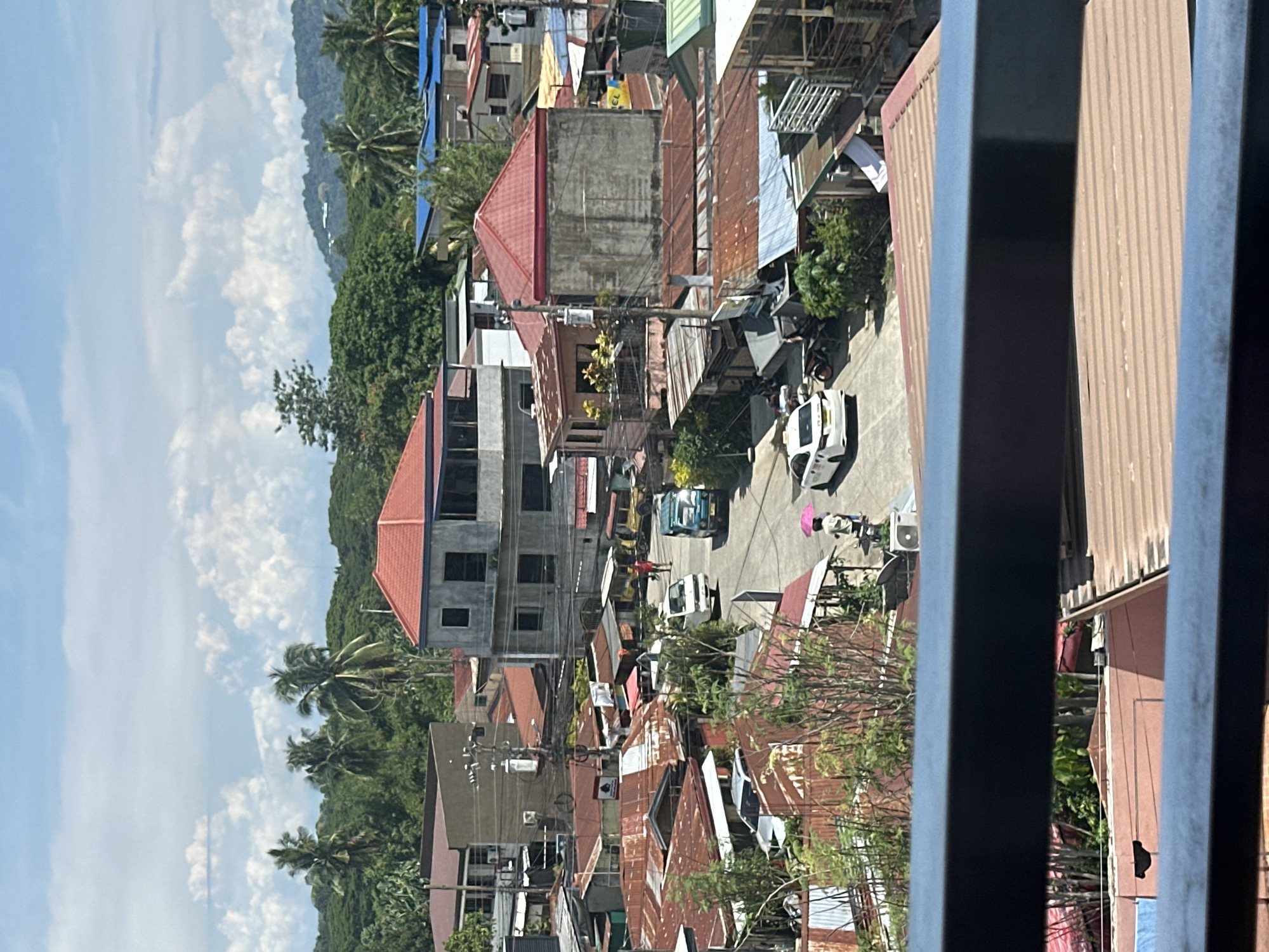 The image shows a densely populated urban area in Davao City, Philippines. The perspective is from an elevated position, likely a balcony or high window, looking down on a street with a mix of residential and commercial buildings. The buildings are diverse, with a mix of concrete structures, some under construction, and older buildings with corrugated metal roofs, many of which are rusty or weathered.
The street appears to have light traffic, with several cars and motorbikes visible. At least one pedestrian with a pink umbrella walks along the side of the road. Power lines are strung haphazardly between the buildings, creating a web of wires overhead.
The surrounding environment includes lush green vegetation, with palm trees and other tropical plants interspersed among the buildings. In the distance, there are hills or mountains covered in dense forest. The sky is partly cloudy, with patches of blue visible. The image is taken during daylight hours, and the lighting is bright.
The composition includes a partial view of the balcony railing, which appears in the foreground on the right side of the image.