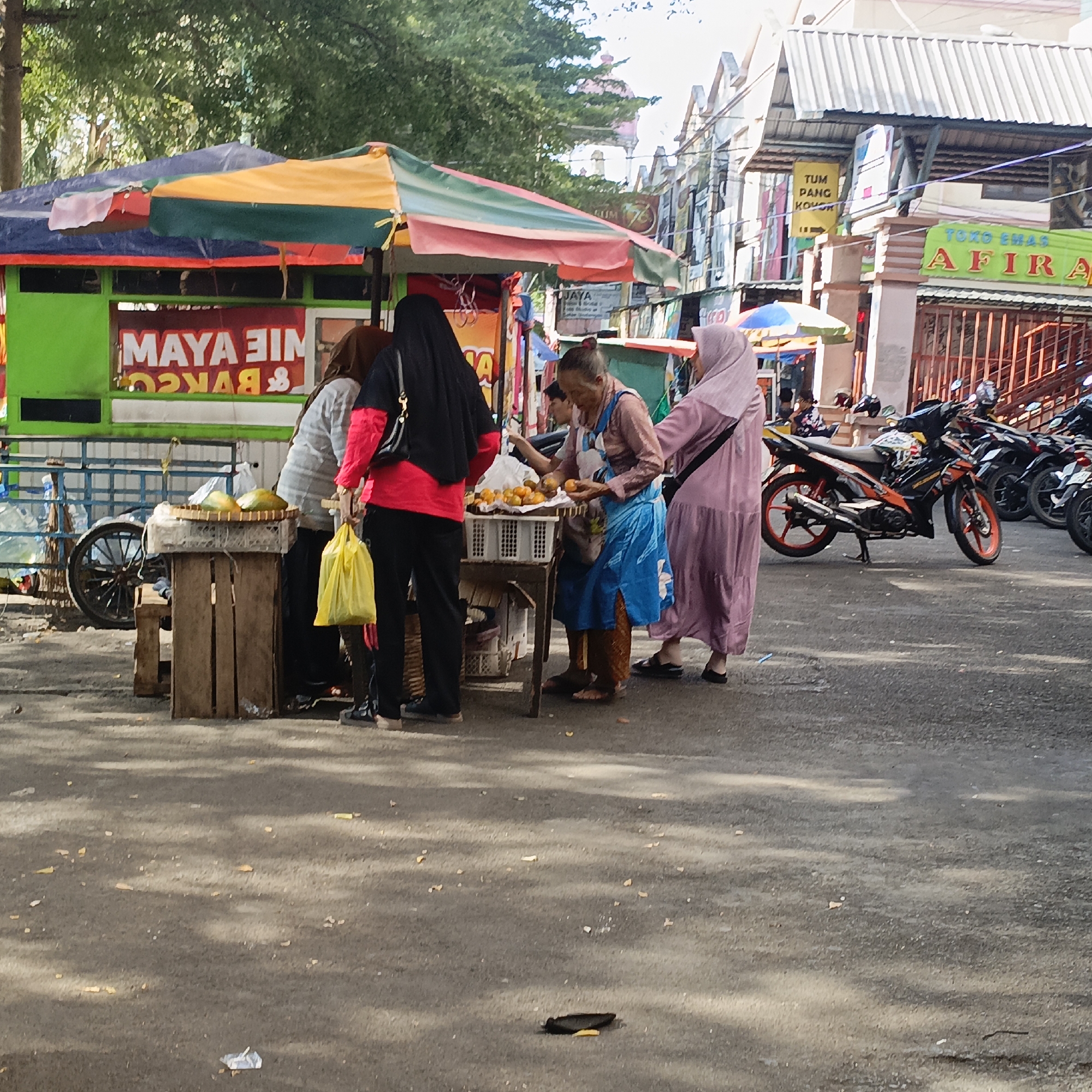 In a street market scene in Salatiga, Indonesia, three women are engaged in transactions at a food stall. The stall, identified by the text "MAYA AIM" and possibly "BAKSO," is covered by a colorful striped awning in shades of green, yellow, and red. A woman in a black hijab and red shirt stands behind the counter, holding a yellow plastic bag. Another woman, wearing a light brown headscarf and a blue apron over an orange shirt and brown patterned skirt, is actively serving food, holding oranges in her hands. A third woman in a light purple hijab and dress is standing on the right, facing the stall. In front of the stall, a wooden stand holds a woven tray with two large green fruits, likely papayas, and a white plastic crate filled with small orange fruits, possibly tangerines. Behind the stall, a blue-wheeled cart is visible.
The surrounding environment suggests a bustling street. Several motorcycles are parked behind the food stall, including a black and orange one with red rims. Buildings line the street in the background, with one featuring a prominent sign that reads "TOKO EMAS AFIRA" (Afira Gold Shop). Another sign above it displays "TUM PANG KOVO". Trees with green foliage are visible overhead. The ground is paved asphalt, with scattered small debris, and shadows suggest a bright, sunny day.