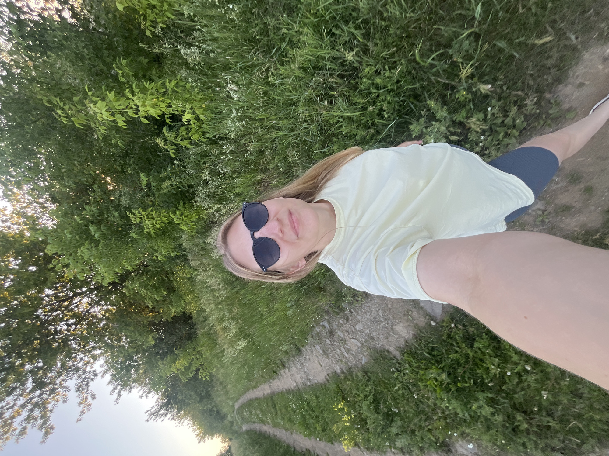 A woman with blonde hair and sunglasses is taking a selfie while standing on a dirt road. She is wearing a pale yellow t-shirt and dark grey shorts. The road is flanked by tall green grass and trees. The sun is setting, casting a warm glow on the scene. The location is described as Бруснятское, Russia.