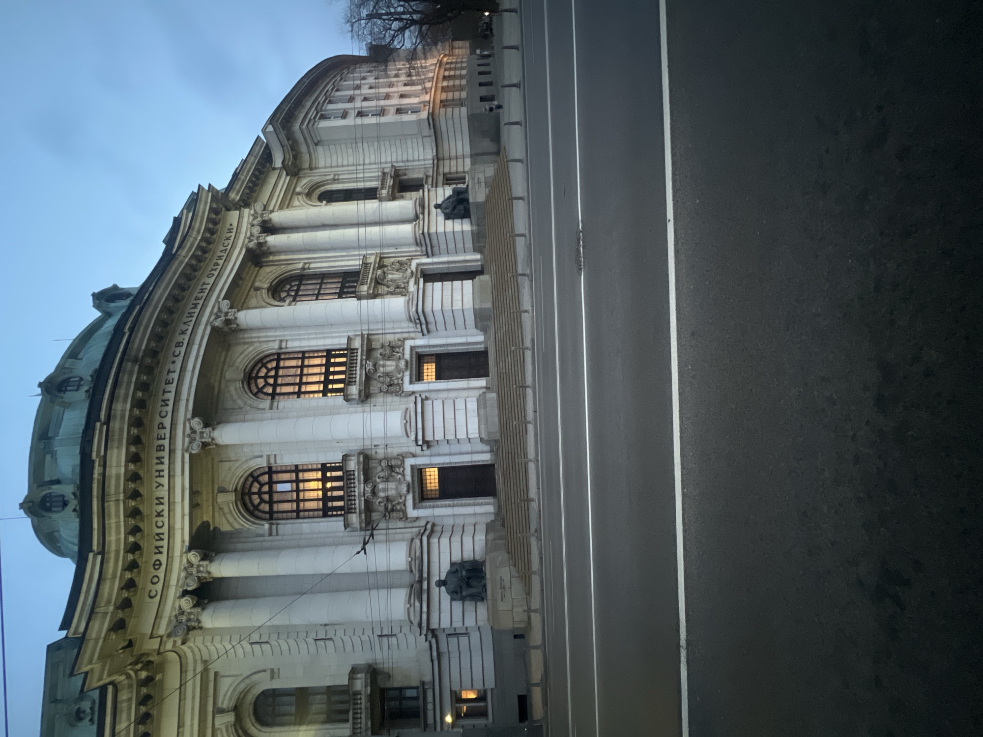 The image depicts the facade of Sofia University "St. Kliment Ohridski" in Sofia, Bulgaria. The classical building features columns, arched windows with light emanating from within, and statues. The inscription "СОФИЙСКИ УНИВЕРСИТЕТ СВ. КЛИМЕНТ ОХРИДСКИ" is visible. A road with white dividing lines runs alongside the building. The sky is overcast, suggesting either dusk or dawn.