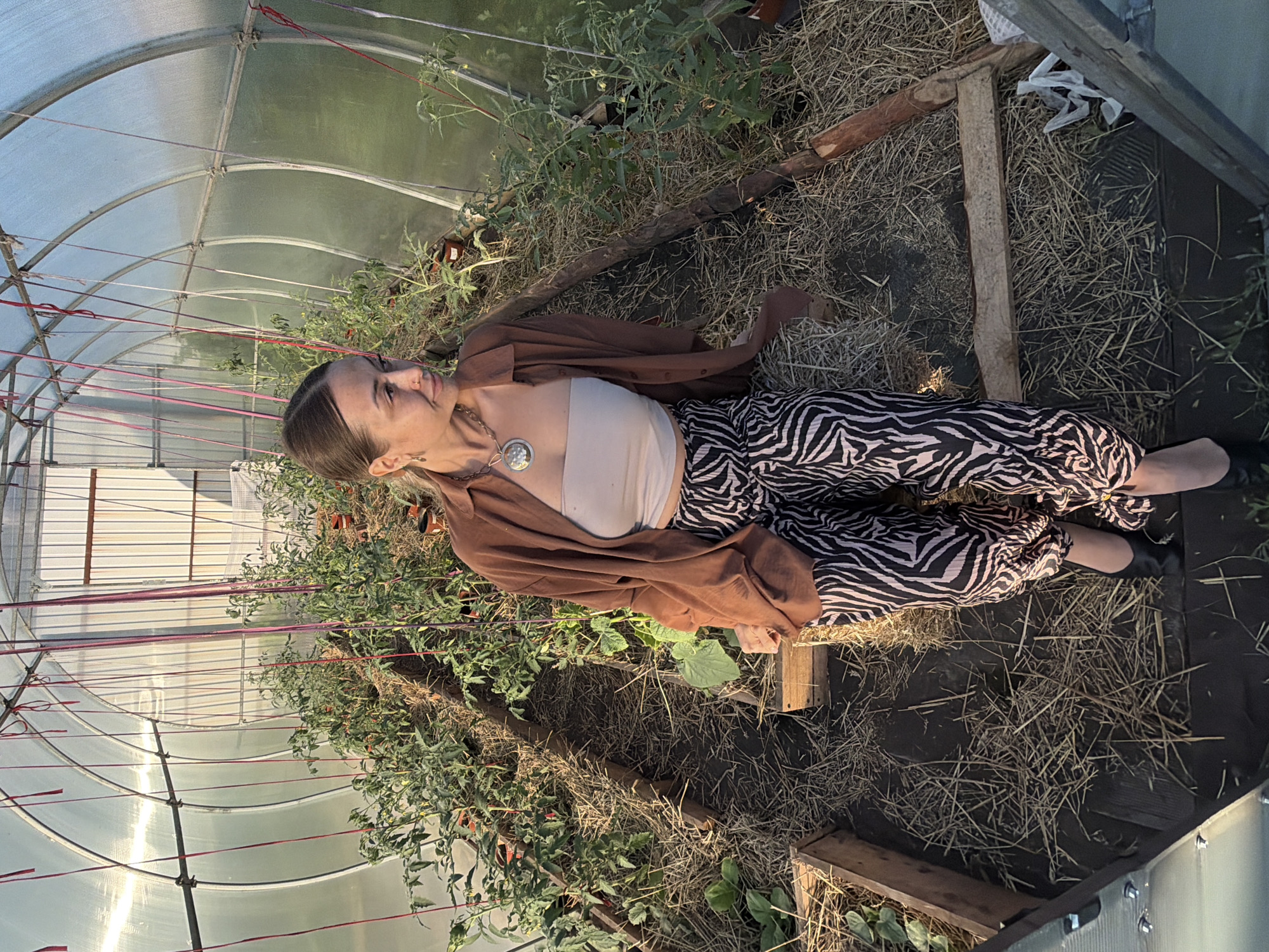 A full shot of a woman with her hair pulled back and wearing a brown button-up shirt and zebra-striped pants sits in a greenhouse. She is looking to the right. Tomato plants are visible in the background, growing in raised beds covered with straw. The greenhouse has a curved, transparent roof supported by metal poles. The ground inside the greenhouse is also covered with straw.