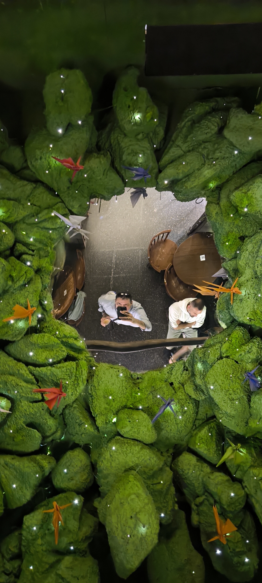 The image shows a reflected view of two men in a restaurant in Barcelona, Spain, using a convex mirror with a textured green frame resembling moss-covered rocks adorned with origami cranes in various colors. The reflection shows the interior of a restaurant with a dark gray floor, round wooden tables, and chairs. One man is holding a phone, seemingly taking a photo, while the other stands beside him. The environment around the mirror gives the impression of a fantastical or nature-themed setting within the restaurant.