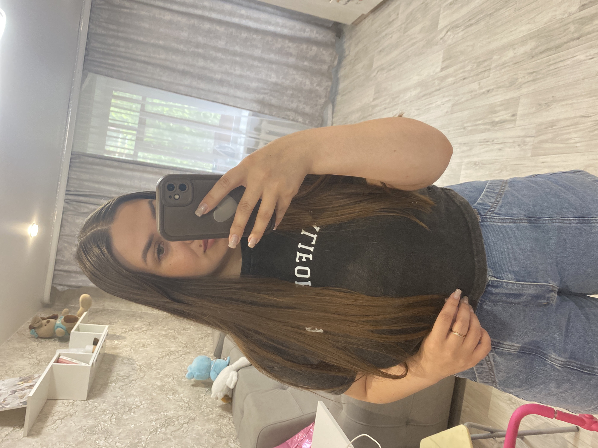 A young woman with long brown hair is taking a selfie in what appears to be an indoor setting. She is wearing a dark t-shirt with a white inscription and blue denim jeans. She is holding a smartphone, likely an iPhone, with a light-colored case. The background includes a window with light filtering through blinds or curtains, a light-colored floor, and elements of a room, including what seems to be a gray couch or chair and a small white shelf unit with various items on it. The environment suggests a domestic setting. The image does not provide enough information to ascertain that this scene is located in the city of Chaikovsky, Russia.