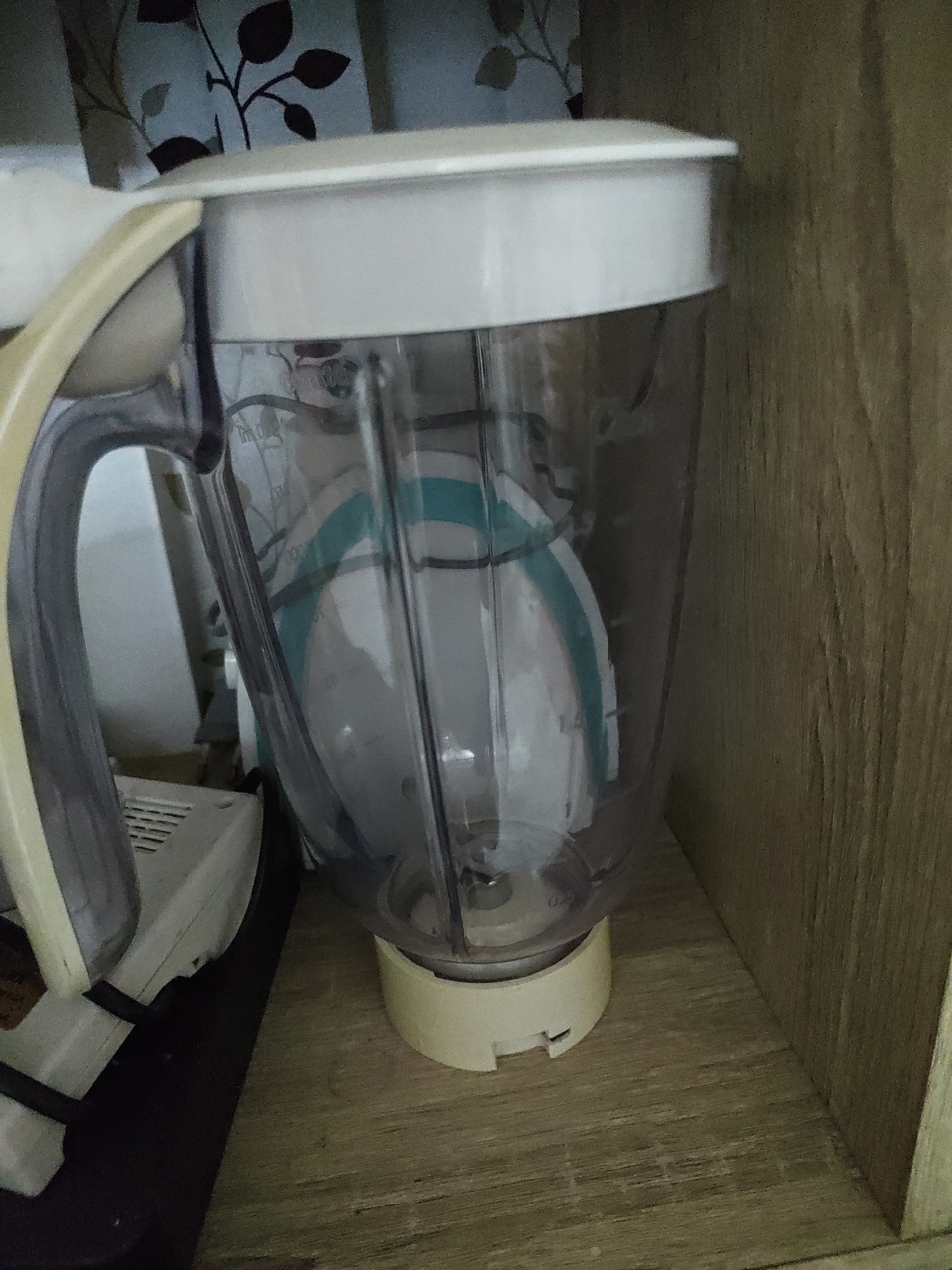 The image shows a close-up of a blender pitcher and its base, which is placed on a wooden shelf inside a cabinet. The blender is white and clear plastic with a grey handle. Inside the pitcher, there appears to be a white plate with teal and blue accents. To the left of the blender, a white appliance with a black cord is visible. The background shows a patterned curtain and the wooden interior of the cabinet. The scene is indoors, and there are no people present. The lighting is dim, suggesting it might be an interior shot taken during the day or in low light conditions. The overall impression is a still life of kitchen appliances stored away. This location is not specified.