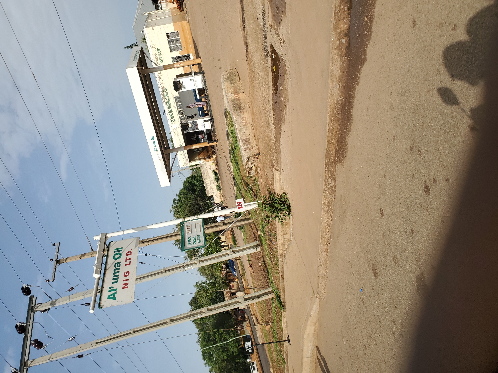 An outdoor daytime medium shot captures a fuel station in Bukuru, Nigeria, under a clear sky with wisps of clouds. Power lines crisscross the frame, supported by tall concrete poles. A prominent sign for "Al'uma Oil NIG LTD." hangs from one of these poles. To the right, the fuel station's white canopy is visible, bearing the "Al'uma Oil" logo and "SUPER MARKET" text. The building behind the canopy is light-colored with yellow accents and has windows. In the foreground, a paved road curves to the left, with a concrete barrier in the middle ground marked with "NO SMOKING". Several vehicles are present: a blue car is parked near the fuel pumps, and other cars are parked in the background. A few individuals are scattered around the station, with one person standing near the fuel pumps and two others in the background. Trees and vegetation surround the area, adding to the natural environment. A small green sign displays prices for "PETROL", "DIESEL", and "KEROSINE". A black sign with white lettering states "YES FUEL".