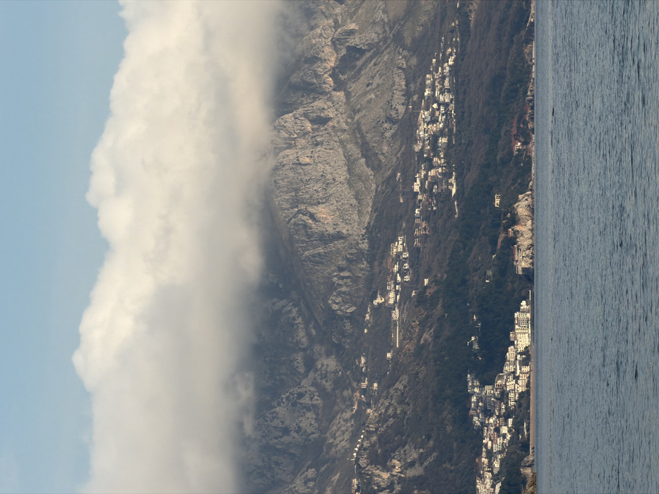This image captures a panoramic view of the Budva Riviera coastline in Montenegro, observed from the Adriatic Sea looking towards the shore. The scene is dominated by a clear separation of three horizontal planes: sea, land, and sky.
The foreground consists of a calm body of water, likely the Adriatic Sea, occupying the bottom quarter of the frame. Its surface is dark blue-grey, exhibiting minor ripples with no discernible vessels or individuals present.
The midground features the verdant, tree-covered slopes of coastal hills that rise steeply from the sea, interspersed with significant residential development. A prominent cluster of white, multi-story buildings with various roof colors lines the coastline on the left, forming a developed resort area such as Bečići or Rafailovići. To the right of this settlement, a distinctive rocky promontory or islet extends into the sea, topped with a cluster of historic-looking, fortified buildings, identifiable as the Sveti Stefan islet. Numerous scattered white and light-colored residential structures are visible on the green-brown slopes, ascending towards the mountains.
The background is dominated by rugged, grey-brown mountains, part of the Dinaric Alps (e.g., Paštrovska Gora or Lovćen range), which rise abruptly behind the coastal settlements. The upper portions of these mountains are partially obscured by a dense band of low-lying, white and grey clouds or mist, which extends across the peaks. Above the cloud layer, a narrow strip of clear, pale blue sky is visible at the top of the image.
The lighting suggests either early morning or late afternoon, with soft, diffused illumination across the landscape. No specific human activities or individuals are discernible due to the long-distance perspective.