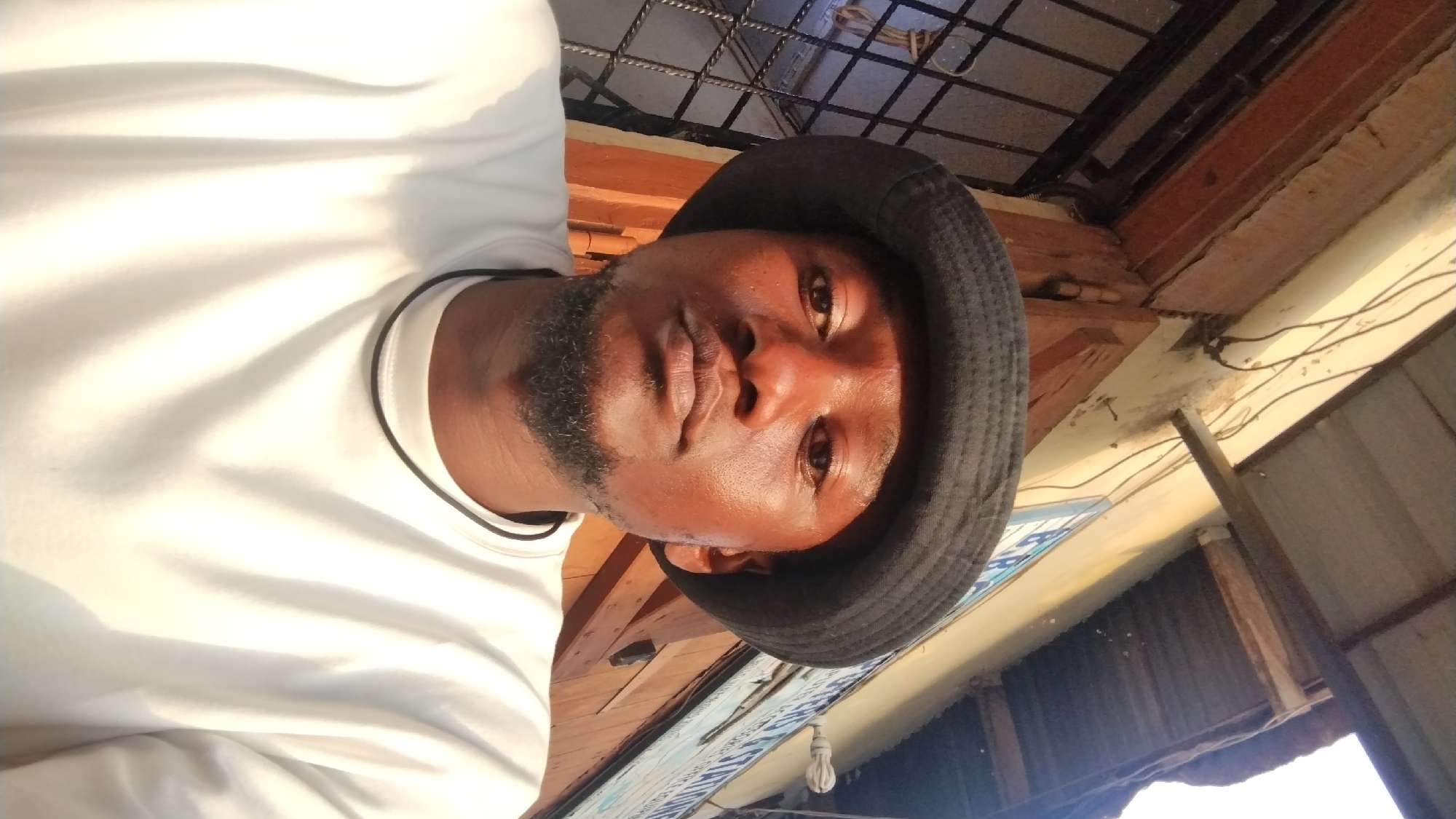 The image shows a man from the chest up, wearing a white t-shirt with a black trim around the neck and a black bucket hat. The man has a beard. The photo appears to be taken from a low angle, looking up.
The background includes glimpses of a ceiling with exposed wooden beams and metal mesh, and what appears to be the exterior of a building. There is a sign with a blue and white design partially visible. Based on the visual elements, I cannot confirm that the photo was taken in the city of Jos, Nigeria.