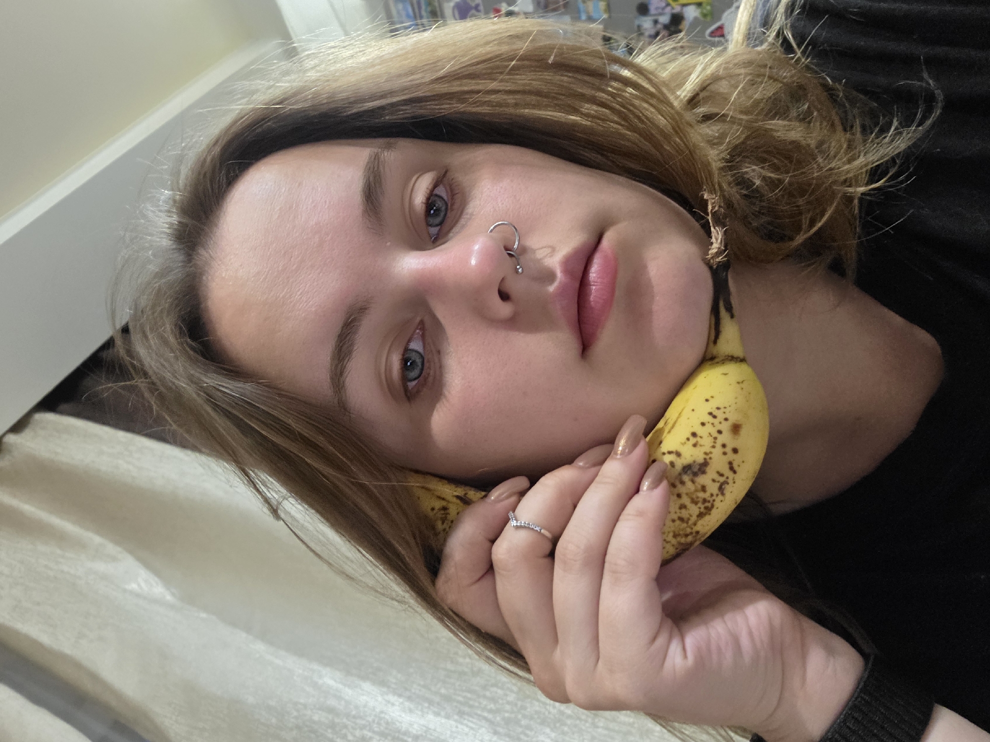 A close-up, over-the-shoulder shot captures a young woman with shoulder-length blonde hair, blue eyes, and a silver nose ring. She is holding a banana to her ear as if making a phone call. Her right hand, adorned with a silver ring, is near her cheek. She is wearing a black top. The background features a light-colored curtain and indistinct objects on shelves. The lighting is dim, and the image quality is slightly grainy.