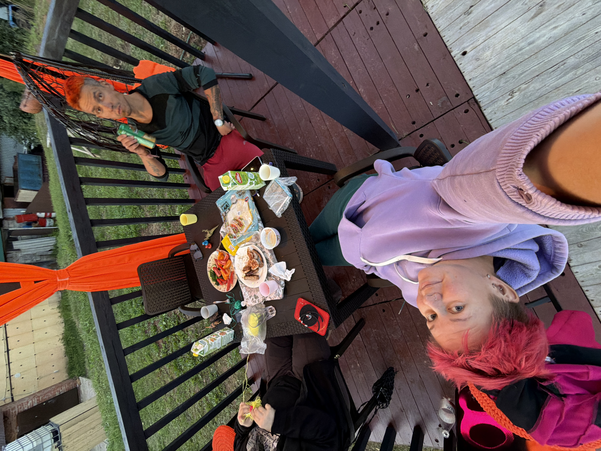 A high-angle, wide shot captures a casual outdoor gathering on a wooden deck, possibly in Chaikovsky, Russia, given the context. Two individuals are visible: one man with bright orange hair is seated in a hanging chair, holding a green can and wearing a dark green and black striped shirt with red shorts. Another person, also with brightly colored pink hair, is in the foreground, leaning back and looking at the camera. A table is laden with food and drinks, including plates of food, a carton of juice, cups, and a phone. The background reveals a grassy area and some utilitarian structures. The overall scene depicts a relaxed, informal social gathering.