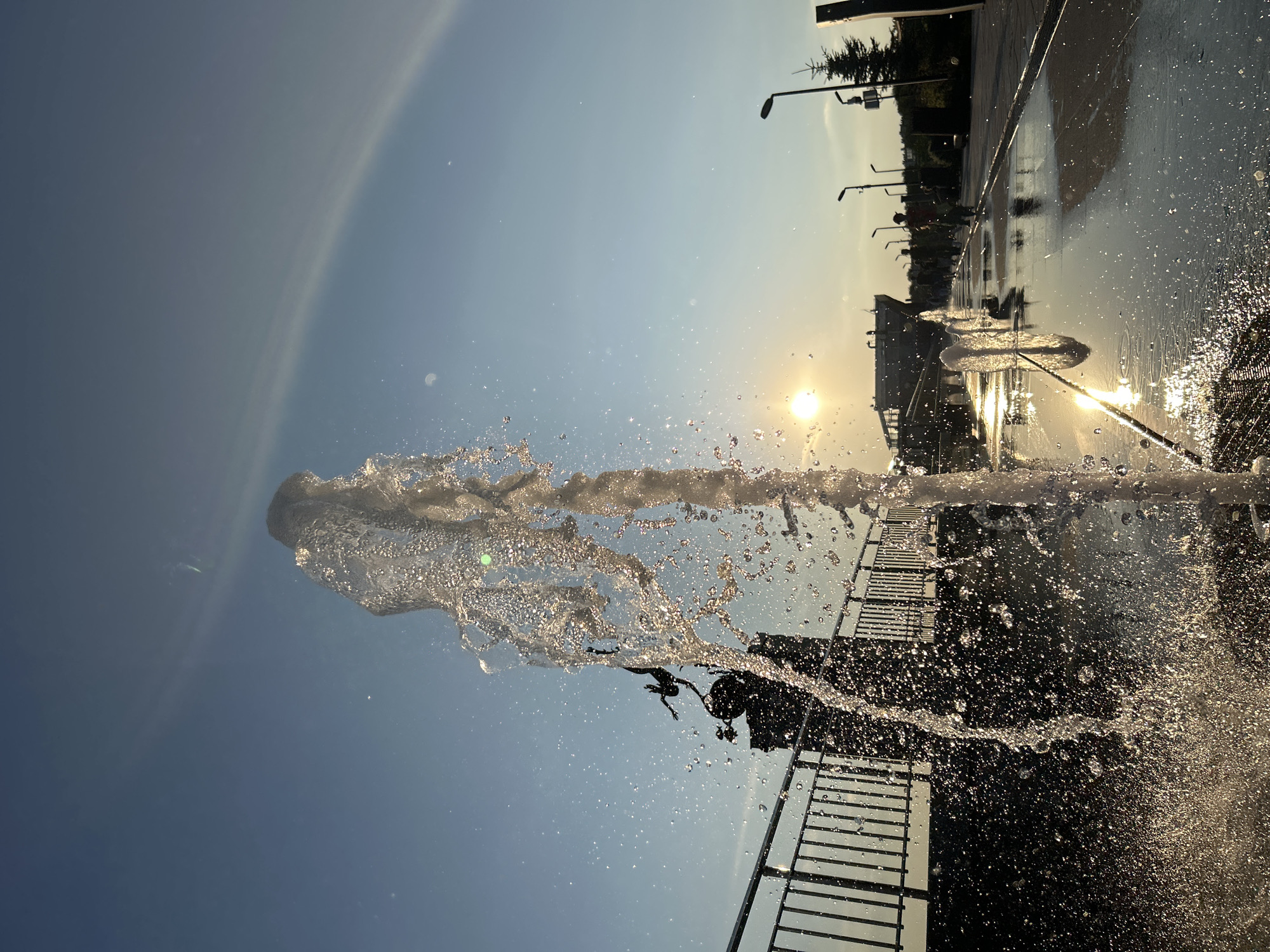The image depicts a sunlit scene with a water fountain in motion, likely in Tolyatti, Russia. The photograph is oriented vertically, with the sky taking up a significant portion of the frame. In the foreground, a jet of water is captured mid-air, spraying droplets in a dynamic arc. The sun is visible as a bright orb in the upper right quadrant, casting a warm glow and creating reflections on the wet ground.
The scene appears to be an outdoor public space, possibly a park or plaza. Hints of this setting include lampposts and a fence-like structure along the right side of the image. The ground is wet and reflective, indicating recent or ongoing activity from the fountain, and perhaps some light rain. Silhouettes of people can be seen in the background, suggesting a leisurely atmosphere. The sky is a clear, pale blue, transitioning to a warmer hue near the horizon, suggesting it is either early morning or late afternoon. There is a faint rainbow or atmospheric optical phenomenon visible in the upper left portion of the sky.
The overall mood is bright and refreshing, with the play of light on water creating a visually engaging scene. The image captures a moment of dynamic natural beauty within an urban environment. No visible text is present in the image.