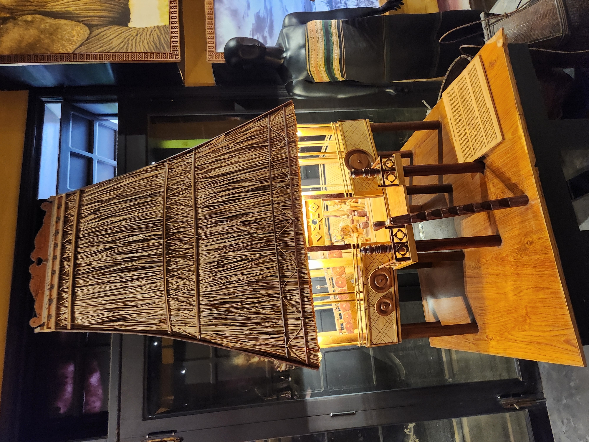 A detailed model of a traditional house, likely from the indigenous cultures of Vietnam, is displayed on a wooden table. The house model features a steep, thatched roof made of dried grass, supported by wooden posts. The interior of the model is illuminated, revealing miniature figures and decorative elements, suggesting a cultural exhibit. To the right of the model, a dark-skinned mannequin is dressed in a traditional woven garment with horizontal stripes of yellow, green, and orange. The mannequin stands beside a woven basket. In the background, a portion of a framed artwork depicting an elephant's trunk is visible on the wall. The scene appears to be an indoor display within a museum or cultural center in Hội An Ward, Vietnam, as indicated by the overall presentation and the nature of the exhibited items.