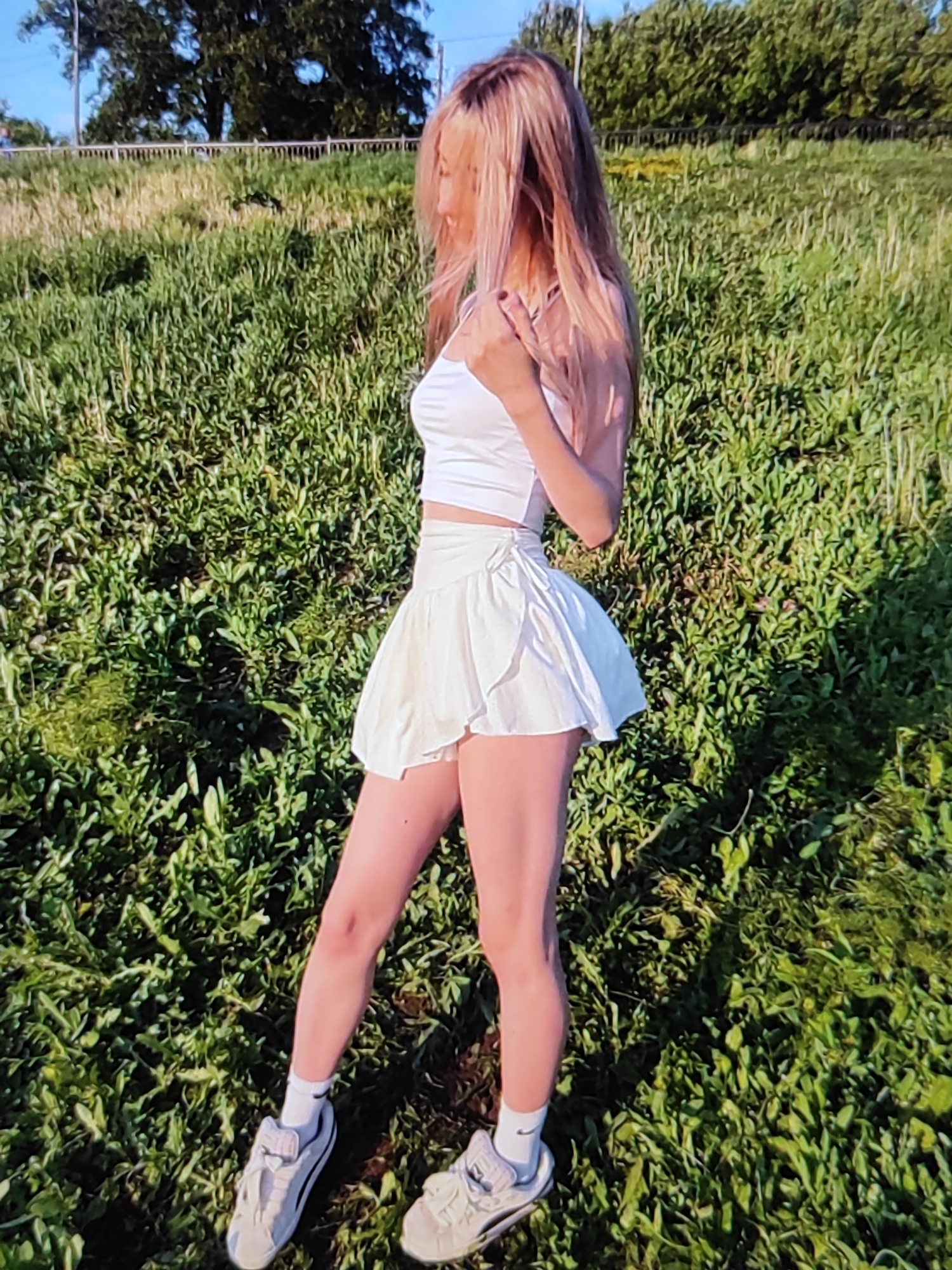 In Fergana City, Uzbekistan, a young woman with long, blonde hair stands in a grassy field under a bright sky. She is wearing a white tank top and a short, white skirt. Her arms are bent, and her hands are near her face, with one finger extended as if gesturing. She is also wearing white sneakers and white socks. The field is covered in green grass, with some shadows indicating sunlight. In the background, there are trees and a fence. The overall scene is outdoors in a natural setting.