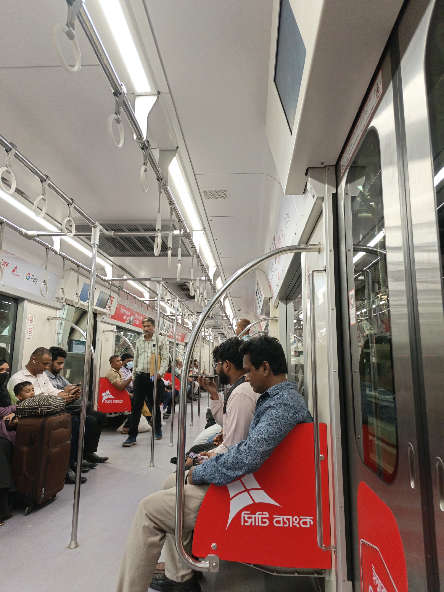 The image shows the interior of a train in Dhaka, Bangladesh. Passengers are seated and standing. One man is standing and appears to be walking through the train. Many people are using their phones. There is luggage on the floor and hanging grips on the ceiling. The seating is red and has text written on it in Bangla.