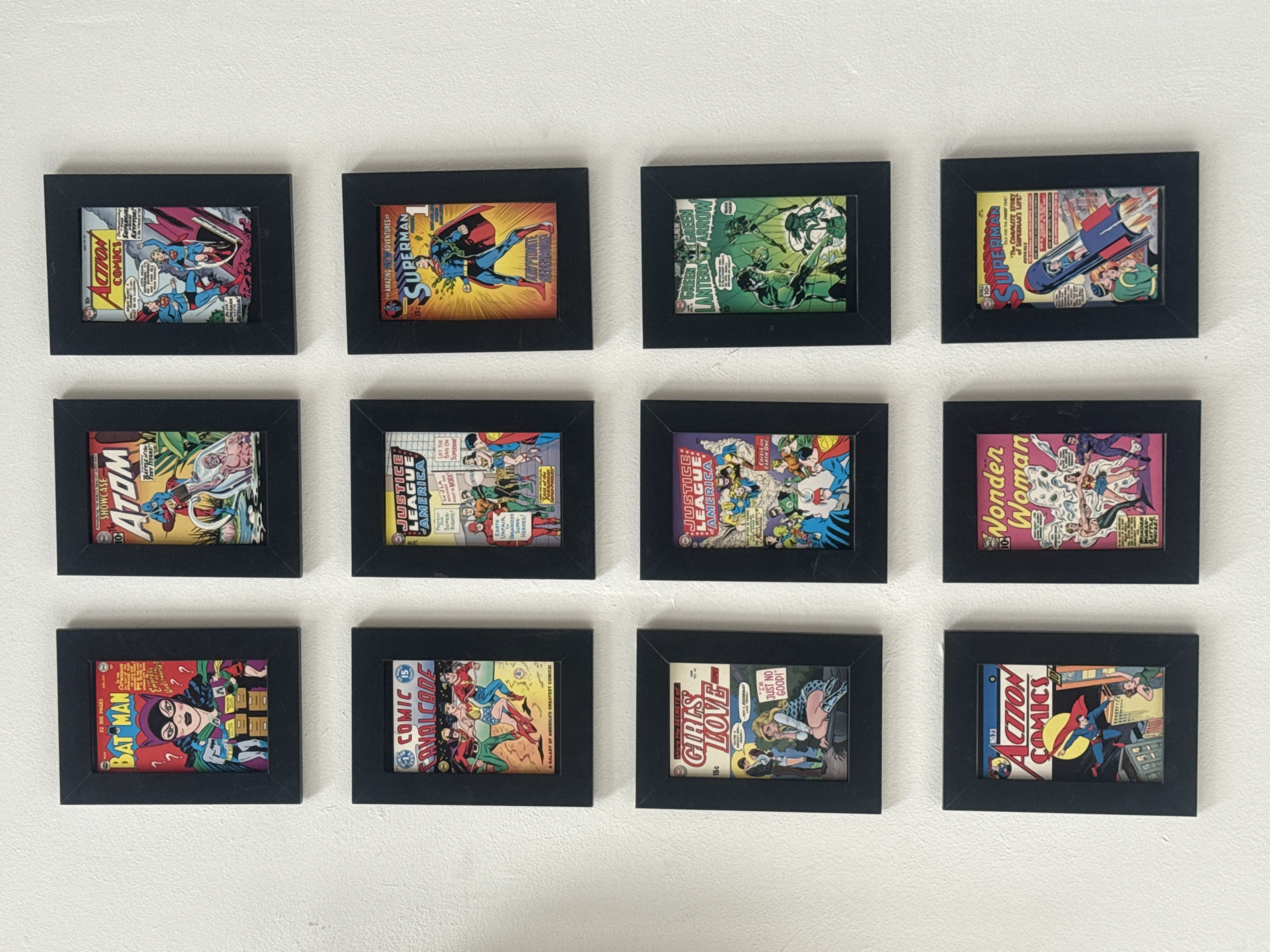 The image shows a collection of twelve framed comic book covers hanging on a white wall. The frames are black and uniformly sized. The comic books are from various publishers and feature different characters including Superman, Batman, Wonder Woman, Green Lantern, and the Justice League of America. The comics appear to be vintage. There are no people visible in the image. The location is not identifiable as Tel-Aviv, Israel based on the image.