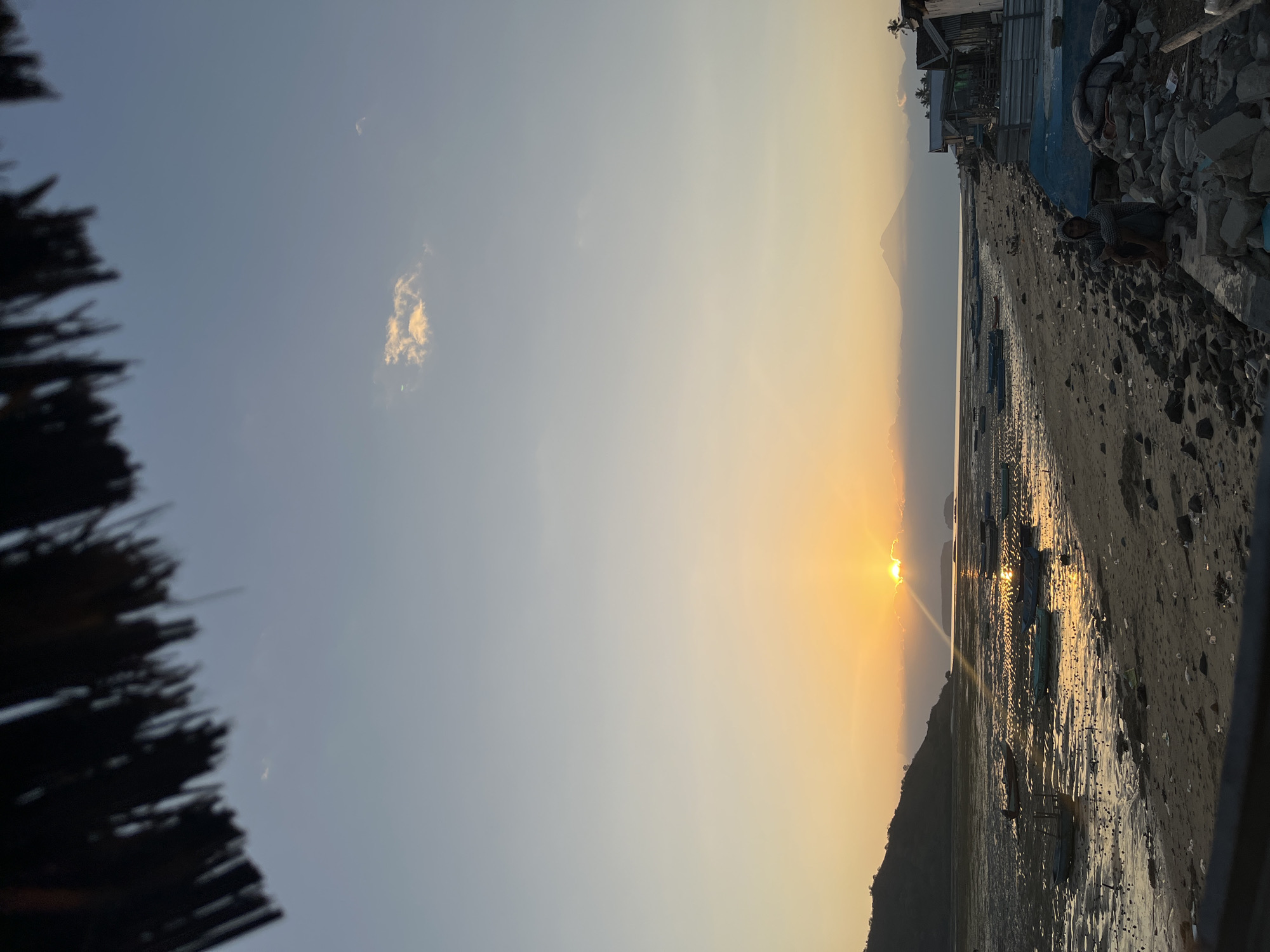 At sunset in Kertasari, Indonesia, a tranquil beach scene unfolds. The sky transitions from a pale blue at the top to a warm, golden hue near the horizon, illuminated by the setting sun peeking through soft clouds. A solitary figure, wearing a hooded garment, sits on a pile of rocks on the sandy shore. In the distance, a majestic, cone-shaped mountain is partially obscured by clouds. Numerous small, blue fishing boats are scattered across the shallow, muddy water and beached on the sand. The foreground is occupied by a dark, thatched roof structure, casting a shadow over the lower portion of the image, and the beach is littered with stones and debris. A small cluster of rustic buildings lines the right side of the scene, suggesting a coastal village.