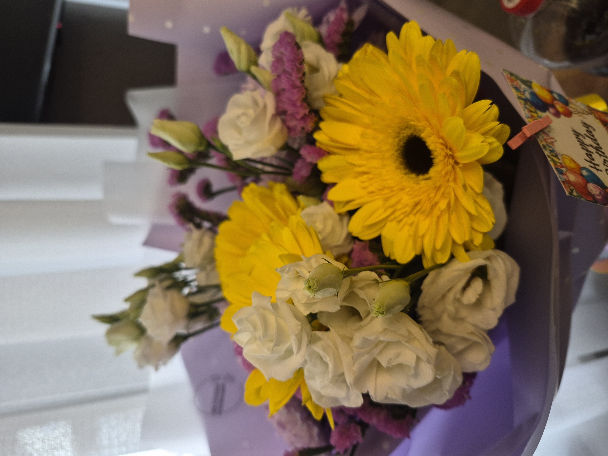This media file captures a vibrant, close-up view of a birthday flower bouquet. The main subjects are a variety of fresh flowers wrapped in purple paper. Dominant in the arrangement are two bright yellow gerbera daisies with distinct dark centers. These are complemented by clusters of delicate white eustoma (lisianthus), some in full bloom and others as closed buds, adding a soft contrast. Sprigs of purple filler flowers, possibly statice, are interspersed throughout, providing texture and depth to the bouquet.
A "Happy Birthday" card is visibly attached to the upper right side of the bouquet, secured by a small pink clothespin. The card's border is adorned with colorful balloon graphics.
The setting appears to be indoors, with the background softly blurred. Hints of white window blinds are visible on the left, suggesting the bouquet might be placed near a window. A faint, out-of-focus object, possibly a bottle with a red cap, can be seen in the upper right. The overall activity implied is the giving or receiving of a celebratory gift. The image conveys a cheerful and appreciative emotion. The location context provided is Chaikovsky, Russia.