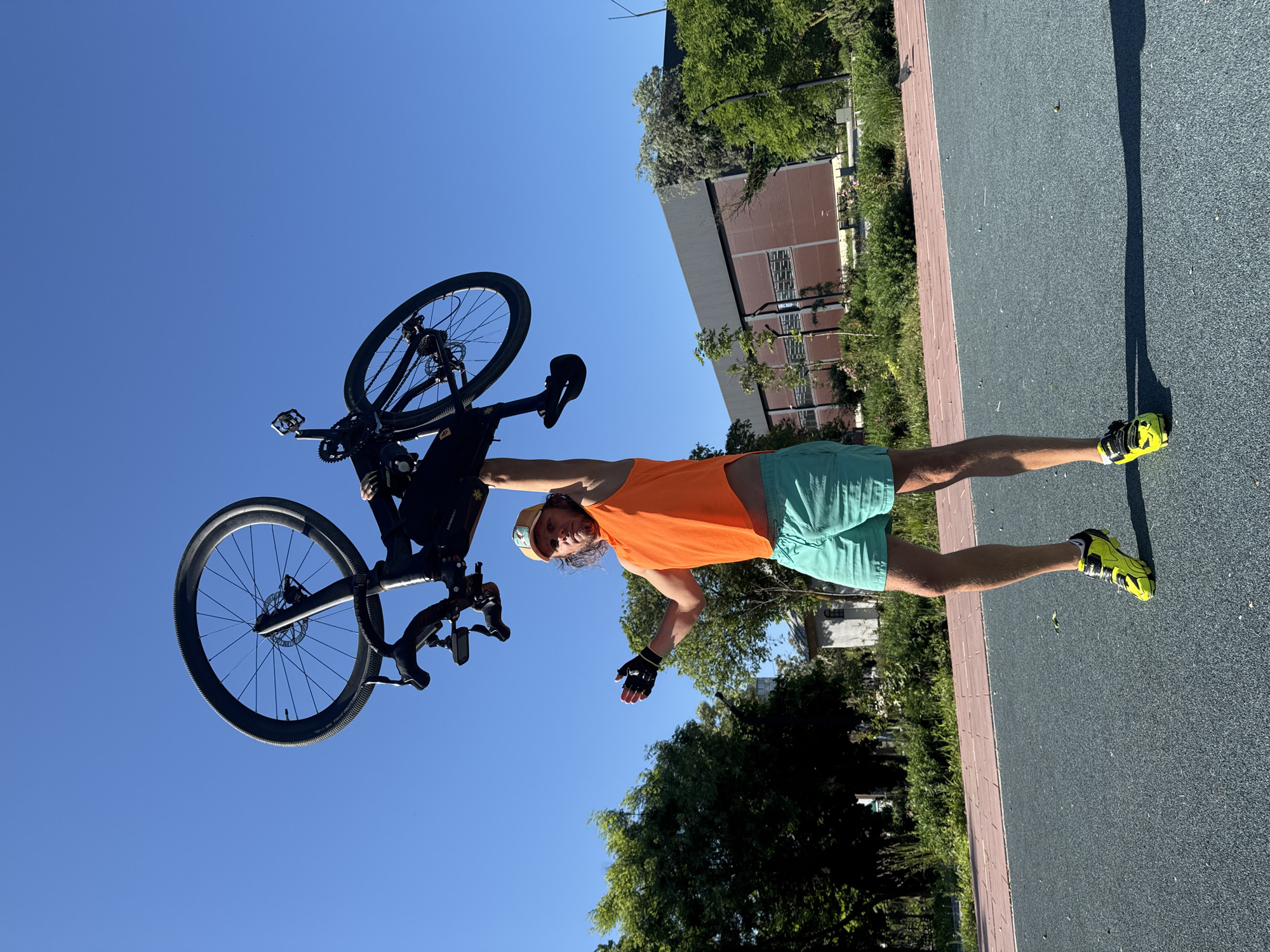 A man in an orange shirt, turquoise shorts, a hat, and yellow shoes is holding a black bicycle in the air. The location appears to be an outdoor area with a paved surface. The sky is blue, and there are trees and a building in the background.