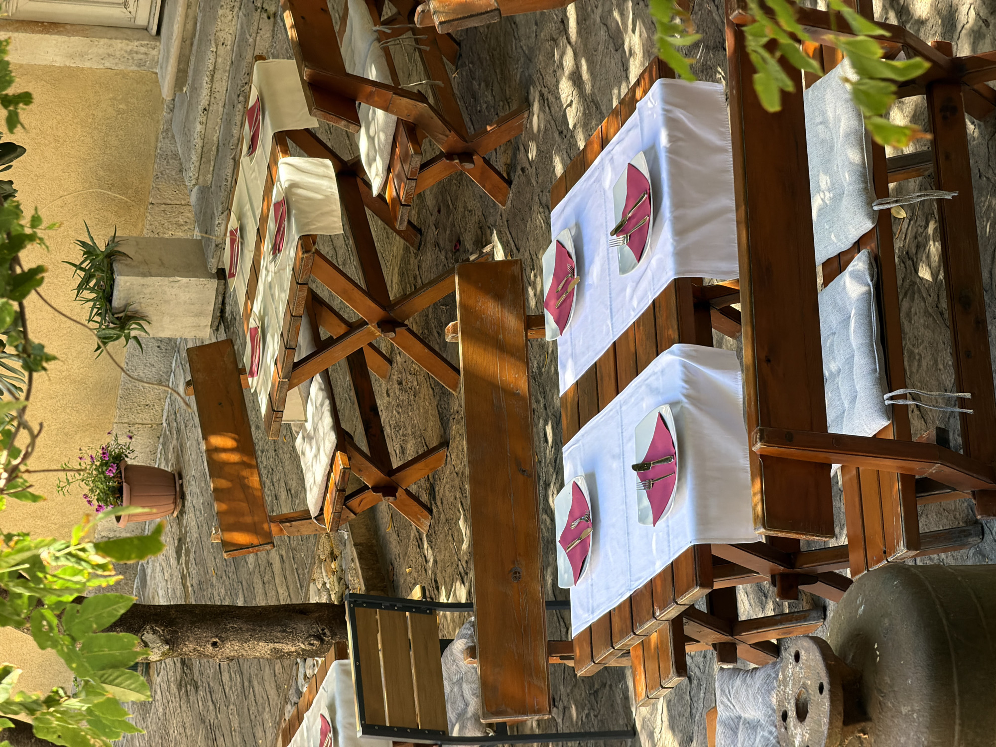 An outdoor dining area with wooden tables and benches is set up on a cobblestone patio. Sunlight filters through the leaves of a nearby tree, casting dappled shadows on the scene. Each table is covered with a white tablecloth and set with a white plate, a pink napkin, and silverware. In the background, there are steps leading up to a building with a yellow facade. Potted plants add a touch of greenery to the setting. The overall impression is one of a relaxed and inviting dining space, likely part of a restaurant or cafe in Budva, Montenegro.