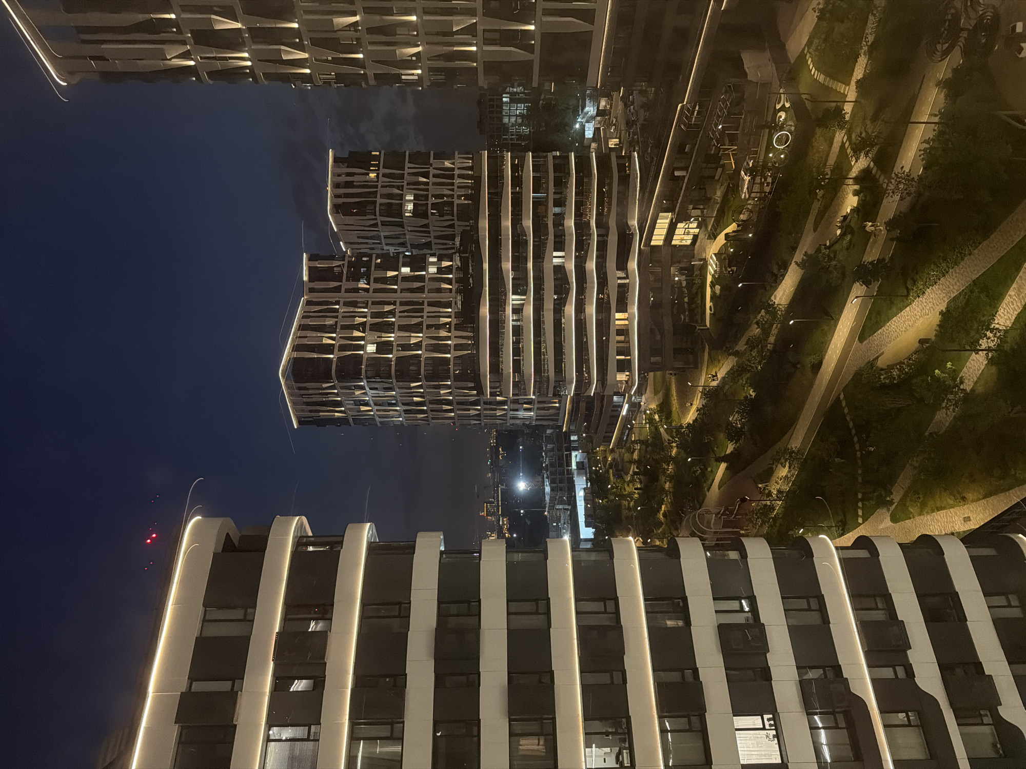 This is an aerial nighttime photograph of a modern residential complex in Moscow, Russia. The complex features several tall, contemporary buildings with illuminated facades, a common architectural style for new developments in the city. The buildings are characterized by their geometric shapes, large windows, and prominent LED lighting that outlines their structures.
In the foreground, a sleek, multi-story residential building is visible on the left, its white horizontal bands and dark window panels illuminated by soft, warm lighting. To the right and further back, other buildings in the complex display similar modern designs, with their facades lit by vertical strips of light.
The scene also includes a well-landscaped urban park or courtyard area situated between the buildings. Winding pathways, illuminated by streetlights, are visible, suggesting pedestrian and cycling routes. Lush green spaces, trees, and modern playground or recreational structures are also part of the park's design. In the distance, the city skyline of Moscow is discernible, with some buildings and lights suggesting a broader urban environment. The sky is dark, indicating it is night, with some subtle cloud formations visible. There are no discernible individuals captured in the scene.