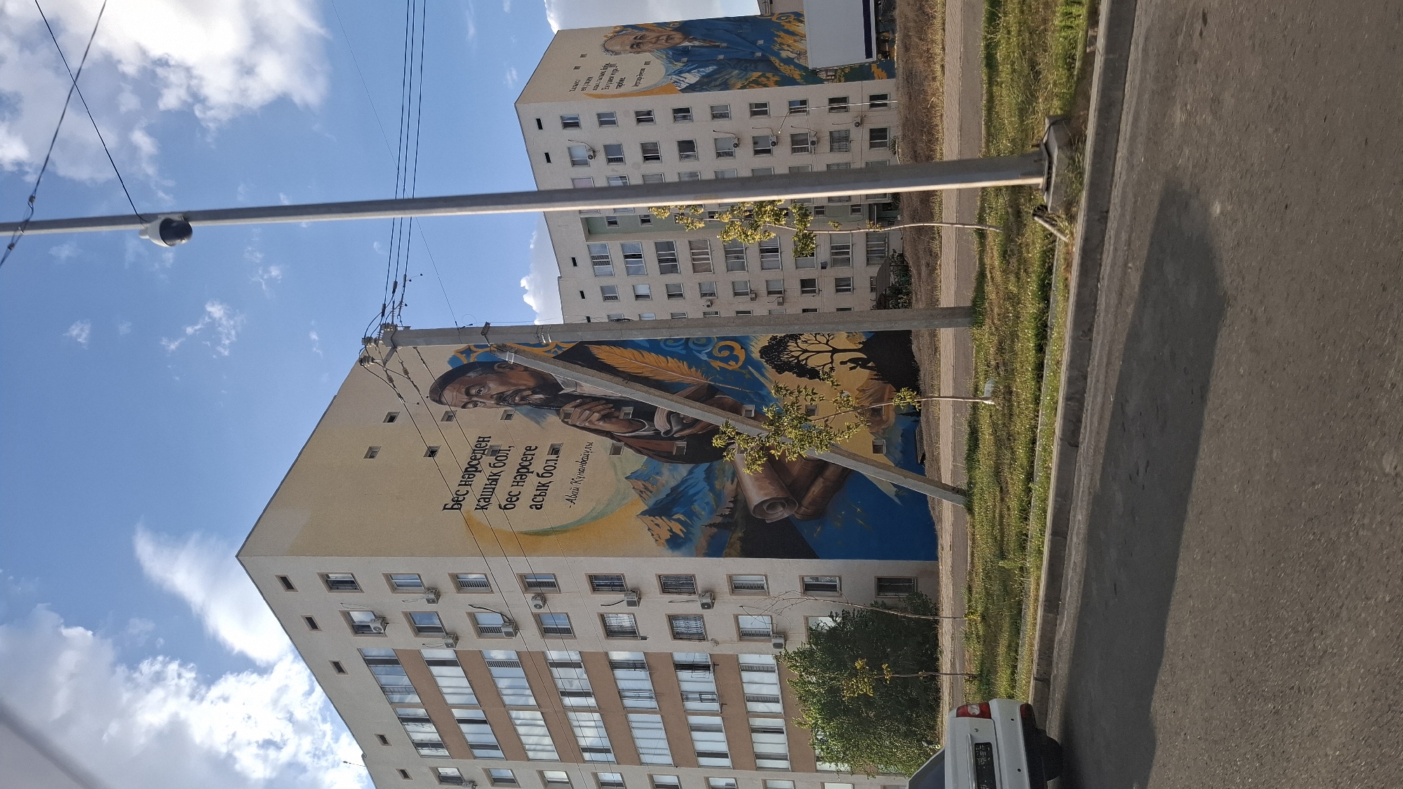 The image depicts a large mural on the side of a multi-story apartment building in Shymkent, Kazakhstan. The mural features a portrait of a man with a beard, wearing a traditional cap, and holding a quill pen. The mural also includes mountainous scenery, a scroll, and a tree with a silhouette of a person beneath it. Text in Kazakh is visible next to the portrait.
To the right, on another apartment building, a second mural is partially visible, featuring a portrait of another man in a suit, with mountainous scenery behind him.
In the foreground, a street runs horizontally across the bottom of the image. A white car is parked on the left side of the street. The environment is sunny with a blue sky and scattered clouds. Streetlights and power lines are also visible in the scene. The buildings appear to be residential apartment complexes. The presence of air conditioning units on the building facades suggests a temperate climate. The mural art indicates a cultural or historical significance to the depicted individuals or themes.