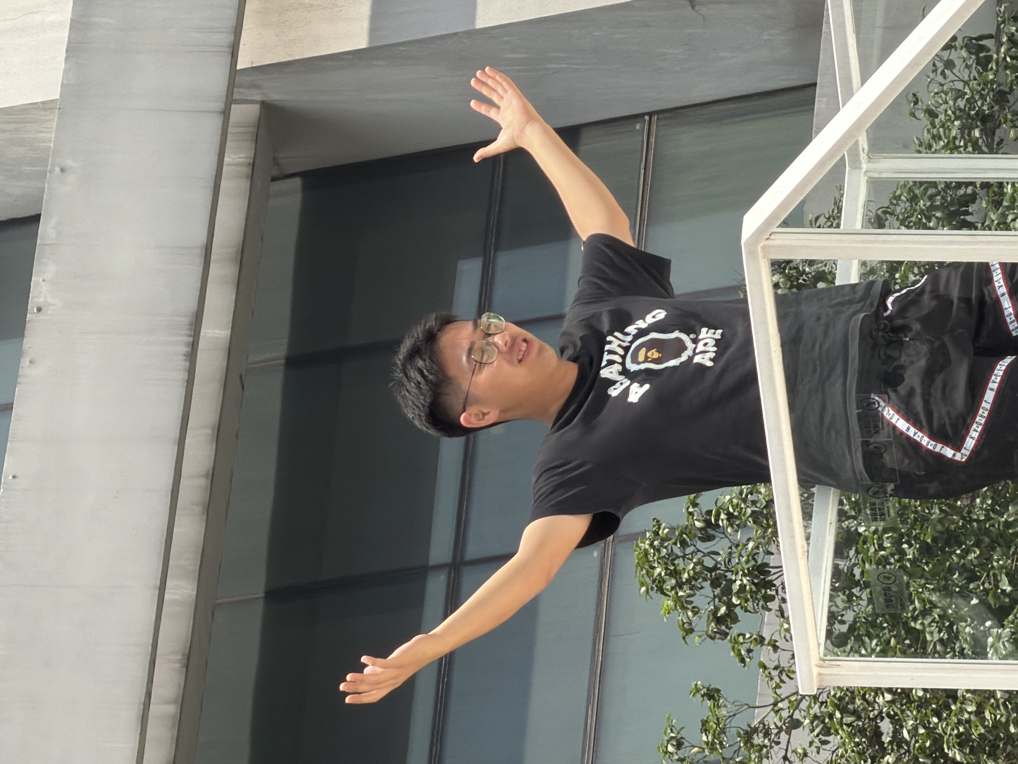 A young East Asian male stands on a white railing, his arms outstretched, facing forward with his head tilted slightly to the right. He wears round, clear-rimmed glasses, a black t-shirt with a white graphic of a monkey face and the word "BAPE" printed on it, and black shorts with red and white stripe detailing down the sides. The background consists of a modern building with large, dark-tinted windows and a light-colored concrete facade. Green foliage is visible behind the railing and the subject. The image appears to be taken outdoors during daylight, likely in a city environment such as Yuzhong District, China, given the architectural style.