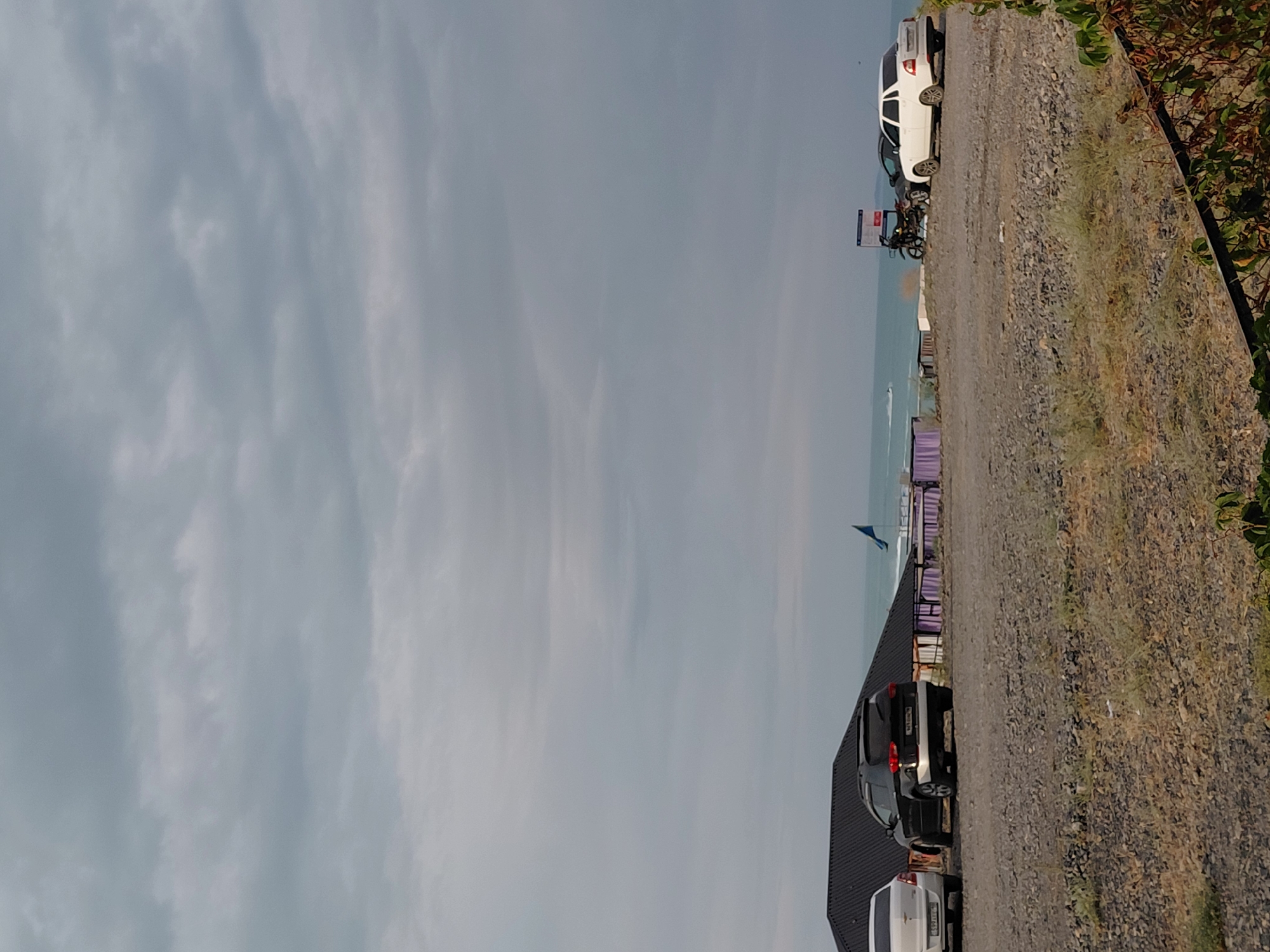 The image captures a cloudy day scene by the sea, likely in Ақши, Kazakhstan. In the foreground, a gravelly, dry area is visible with some sparse vegetation. A dark SUV is parked on the left side, and a lighter-colored sedan is parked to its right. Behind these vehicles, a building with a dark, sloped roof is partially visible. Further back, a blue flag flutters against the backdrop of the sea. The sea itself appears calm, with a jet ski in the distance. On the right side of the image, two white signs with red accents stand upright. A white sedan is parked next to these signs, and a motorcycle is positioned between the signs and a dark car that is mostly obscured. The sky is overcast with numerous clouds, casting a diffused light over the scene.