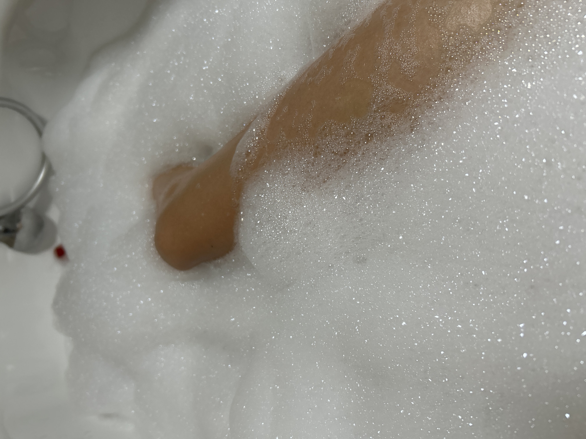 A close-up shot shows a person's leg submerged in a bathtub filled with white foam. The leg appears to be tanned and is partially visible through the bubbles. The foam is abundant and textured, with many small bubbles creating a soft, airy surface. In the upper right portion of the image, a chrome faucet and a small red object are partially visible, suggesting the presence of a bathtub fixture. The overall environment appears to be a bathroom. The location in the city of Vyshevychi, Ukraine, is not depicted or implied by the visual information in the image.