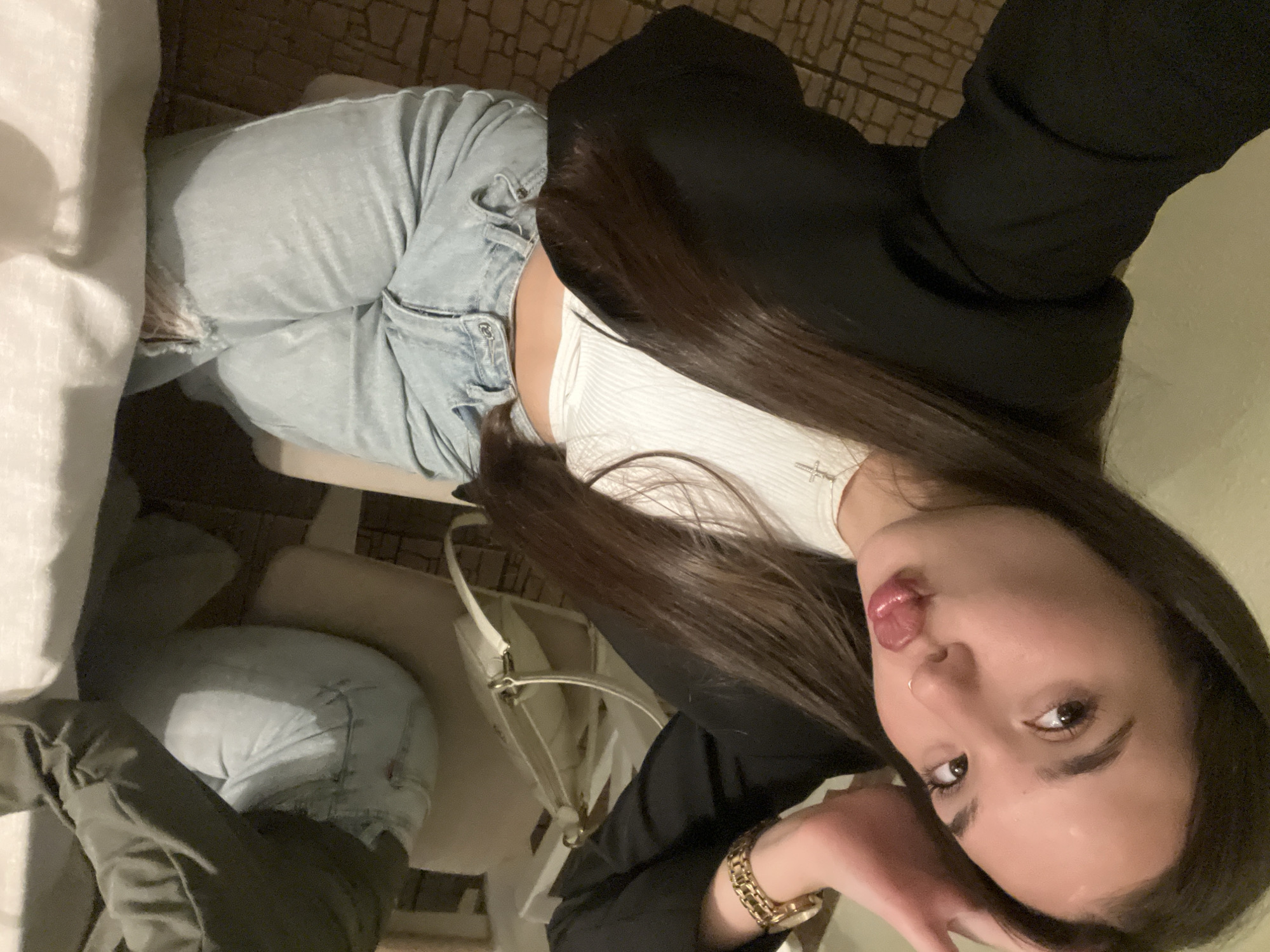 A woman is taking a selfie indoors. She is wearing light blue jeans, a white tank top, and a black blazer. A gold watch is visible on her wrist. A white purse rests on a chair next to her. The location cannot be determined as being in the city of Chaikovsky, Russia, as there is no information available in the image.