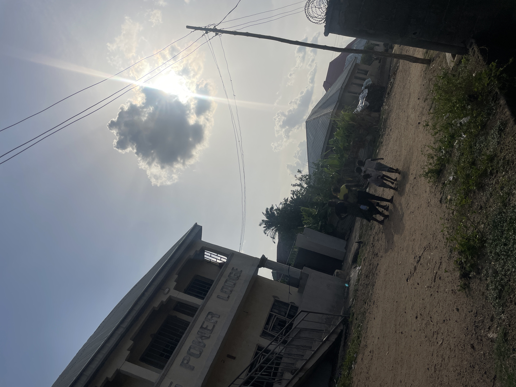This image captures a bright, sunny outdoor scene, likely a street or dirt road, consistent with a semi-urban environment in Lokoja, Nigeria. The sky is a dominant feature, filled with bright sunlight and scattered clouds, creating strong backlighting and a visible lens flare. A wooden utility pole with several power lines stretches across the upper part of the frame.
The ground consists of an unpaved dirt road, which appears to extend into the distance. Sparse green vegetation and bushes line the edges of the road, particularly on the right side. In the mid-ground, a small group of at least three individuals, possibly children or young adults, walk along the dirt road away from the viewer. They seem to be wearing light-colored tops, potentially school uniforms.
On the left side of the scene, a multi-story concrete building stands prominently. The words "POWER LODGE" are visibly embossed on its facade. The building features windows with security bars and an external metal staircase. Further in the background, another building with a dark-colored roof is partially visible, and some tropical foliage, including a palm tree, can be seen behind the structures. Barbed wire or razor wire is present on top of a wall in the upper right background, suggesting a security feature. The scene depicts an ordinary day, likely late morning or early afternoon given the high sun.