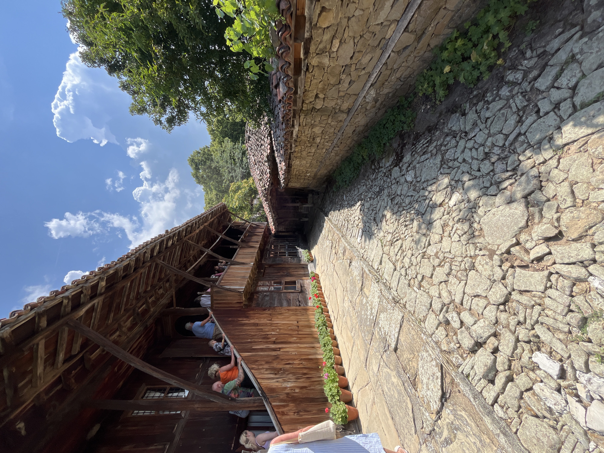 In Zheravna, Bulgaria, a cobblestone path leads past a traditional wooden house with a covered balcony. The house has wooden walls and a tiled roof. Several people are on the balcony and the steps leading up to it. Flower pots with red flowers are lined up along the path in front of the house. To the right of the path is a stone wall with a tiled roof on top. Trees are visible in the background. The sky is blue with white clouds.