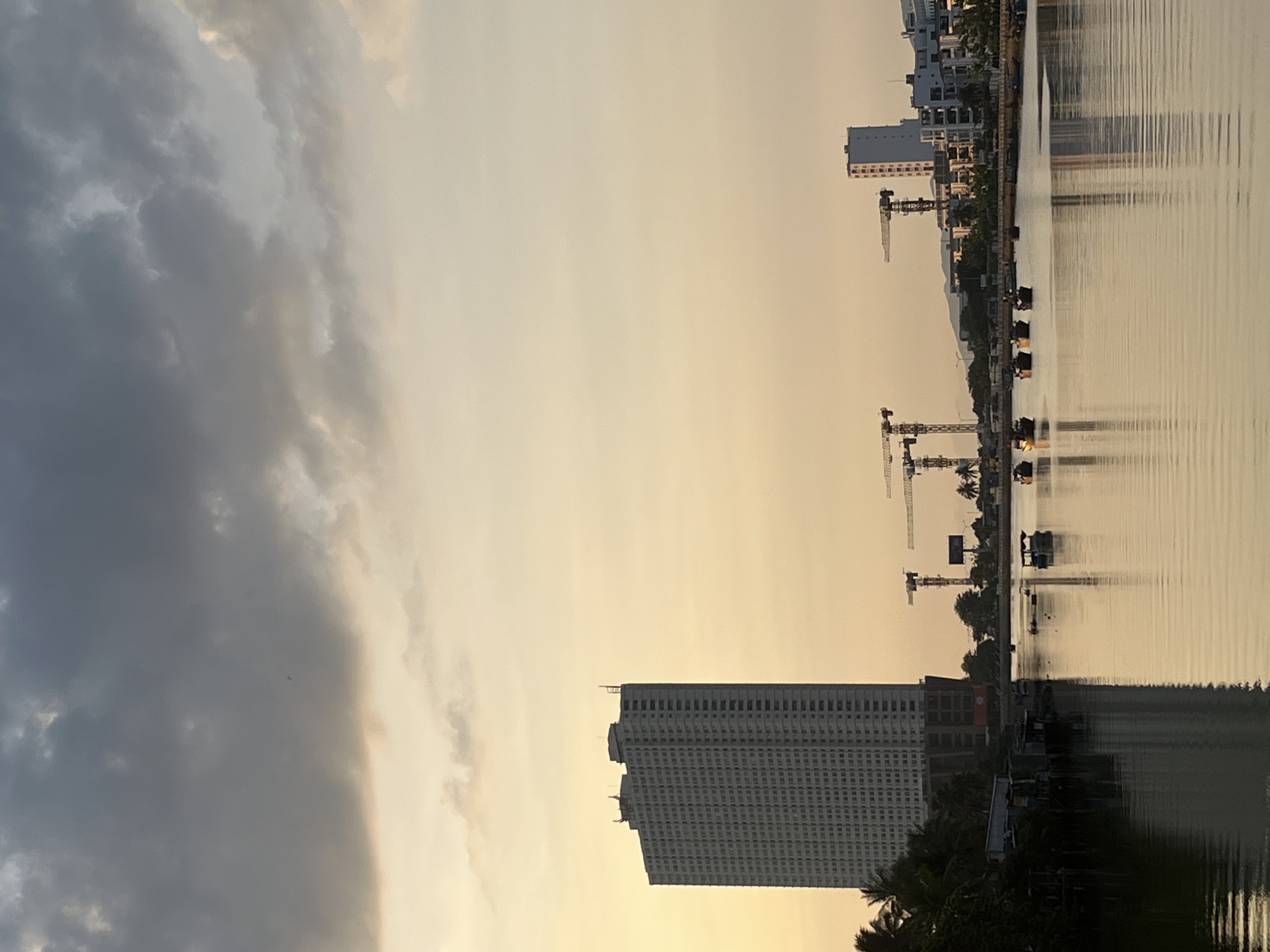 The image shows a view of Nha Trang Ward, Vietnam. The scene is dominated by a large body of water, possibly the Cai River, reflecting the skyline. There are numerous construction cranes visible, indicating ongoing development. Buildings of varying heights are present along the waterfront, including a tall, modern apartment or office tower. The sky is divided between ominous dark clouds on the left side and a golden, hazy sky on the right, which could indicate dusk or sunrise. The presence of trees and foliage suggests some natural elements interspersed with the urban landscape.