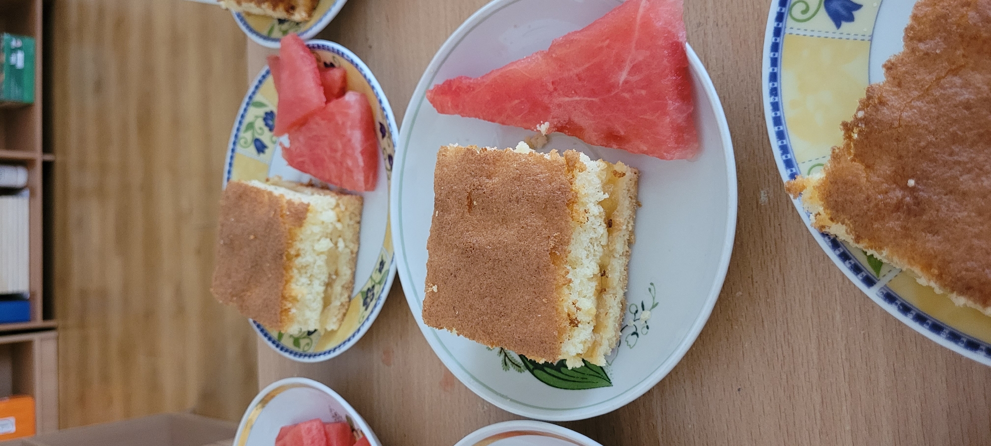 The image shows three plates with slices of cake and watermelon. Two plates are on a wooden surface. One plate in the foreground has a slice of cake with a filling and a slice of watermelon next to it. The plate has a floral design. Another plate in the middle ground, also on the wooden surface, has a slice of cake and several slices of watermelon. A third plate, partially visible on the left side, contains slices of watermelon. In the background, a bookshelf is visible with some items on it, and a wooden floor. The scene appears to be indoors.