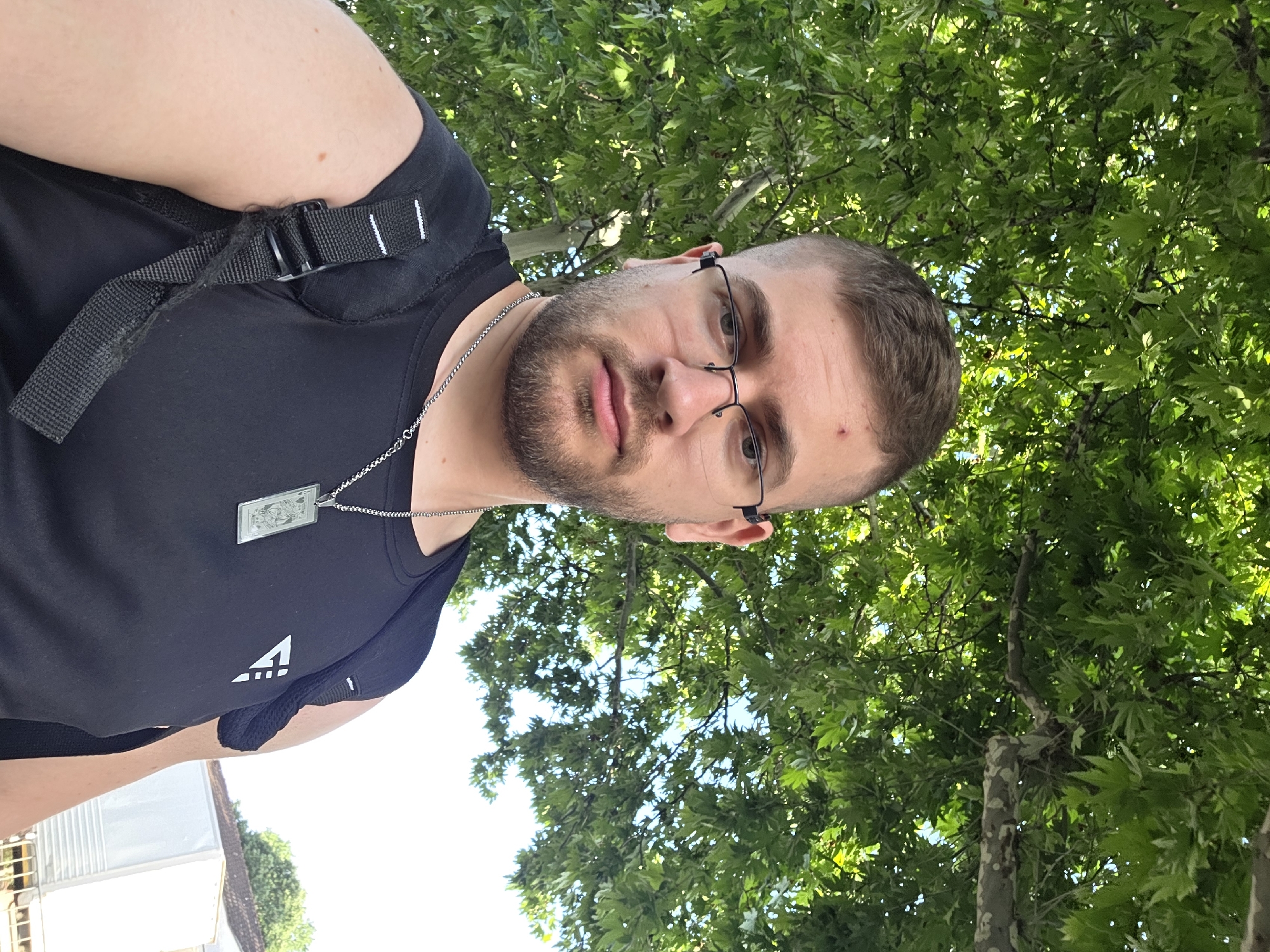 A medium-angle, vertically-oriented shot depicts a Caucasian male with a beard and glasses, positioned in the foreground. He wears a black t-shirt, a silver necklace with a rectangular pendant, and a black backpack over his shoulder. He has a small reddish mark on his left temple.
The background consists of a dense canopy of green leaves from several trees, creating an impression of a wooded area. The sky is visible in small patches through the leaves. In the lower left corner, there is a partial view of a white structure, possibly a building, under bright sunlight.
Given the presence of distinctive plane trees, this scene appears to be located in the city of Plovdiv, Bulgaria, a location known for its plane trees. However, precise geo-location within the city is not discernible from the image alone.