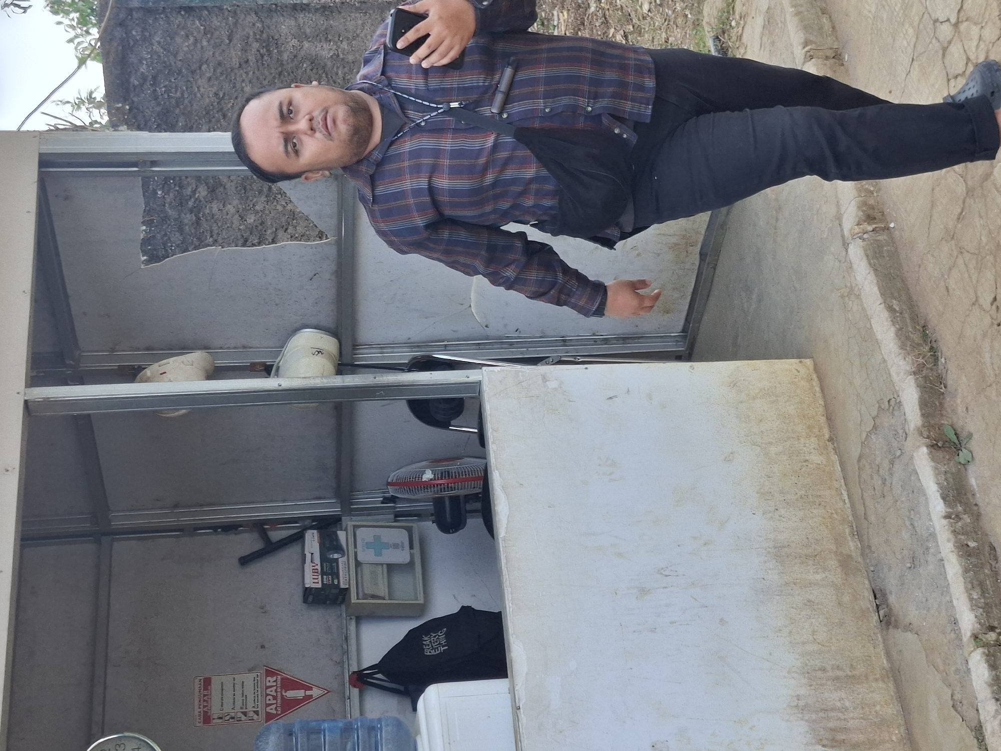 A medium shot shows a man walking towards the right of the frame in front of an open, dilapidated kiosk. He is wearing a plaid long-sleeved shirt, dark pants, and dark slip-on shoes. A black bag is slung across his chest. He is holding a dark smartphone in his right hand. The kiosk behind him appears to be constructed of metal framing and has a broken or missing panel on its right side, revealing a textured wall behind. Inside the kiosk, a blue water jug is visible on the left, along with a black backpack hanging from a hook. A small fan, a first-aid kit in a wooden frame, and a box labeled "LUBNY" are also present. The ground in front of the kiosk is paved with concrete and appears worn and cracked. The environment suggests an outdoor urban setting, possibly in Ciheulang Tonggoh, Indonesia, given the background of a rough, textured wall and some foliage.