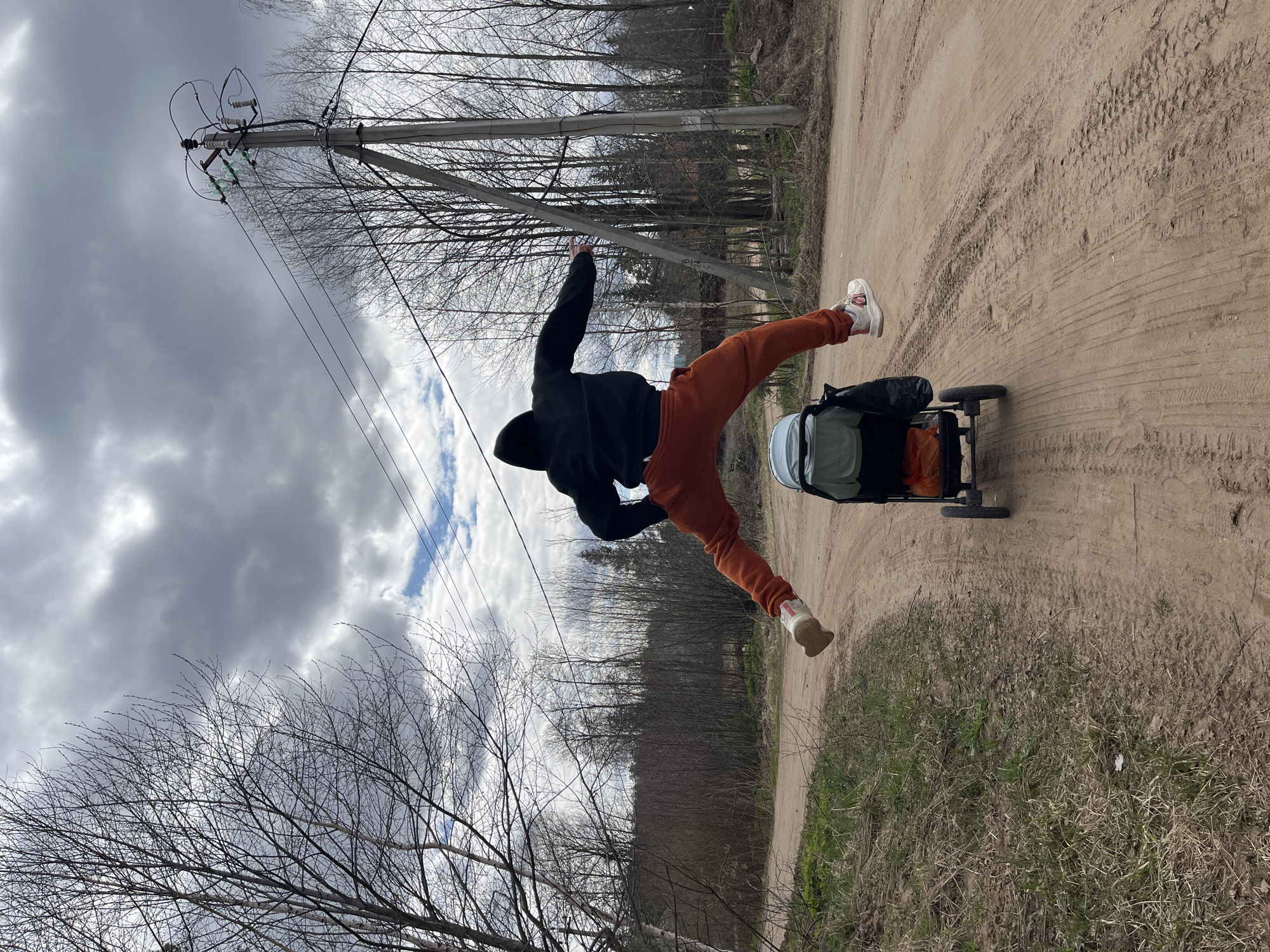 A photograph shows a person suspended in mid-air next to a stroller on a sandy path. The individual is wearing a black hooded sweatshirt and orange sweatpants, with white sneakers on their feet. Their body is oriented at an angle, appearing as if they are jumping or performing a dynamic movement. Beside them is a stroller containing luggage.
The background reveals a dirt path bordered by grassy vegetation on one side. Power lines and a utility pole are visible on the other side of the road. Bare trees line the area beyond the path, hinting at a location near a wooded area. The sky is overcast with clouds and patches of blue.
Based on the metadata, the photograph was taken in Chaikovsky, Russia, on April 16, 2023.