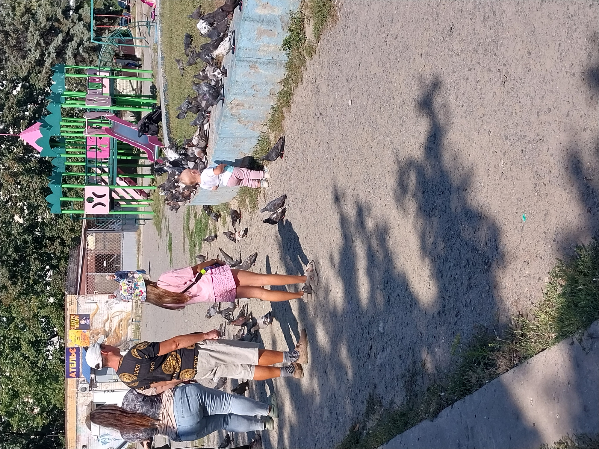 The image depicts a scene on a sunny day in a public outdoor area, likely a park or playground, in Dnipro, Ukraine. In the foreground, a person in light-colored shorts and socks stands facing away from the camera, with a shadow cast on the ground. To the left, a woman in blue jeans and a patterned top is also seen from the back. A young child in a white shirt and pink pants stands facing a flock of pigeons gathered on a low concrete barrier. Behind the child and pigeons, a colorful playground structure with slides and swings is visible. In the background, trees and a building with signage, one sign in Cyrillic, are present. The ground is a mixture of asphalt and dirt. The overall atmosphere is casual and everyday.